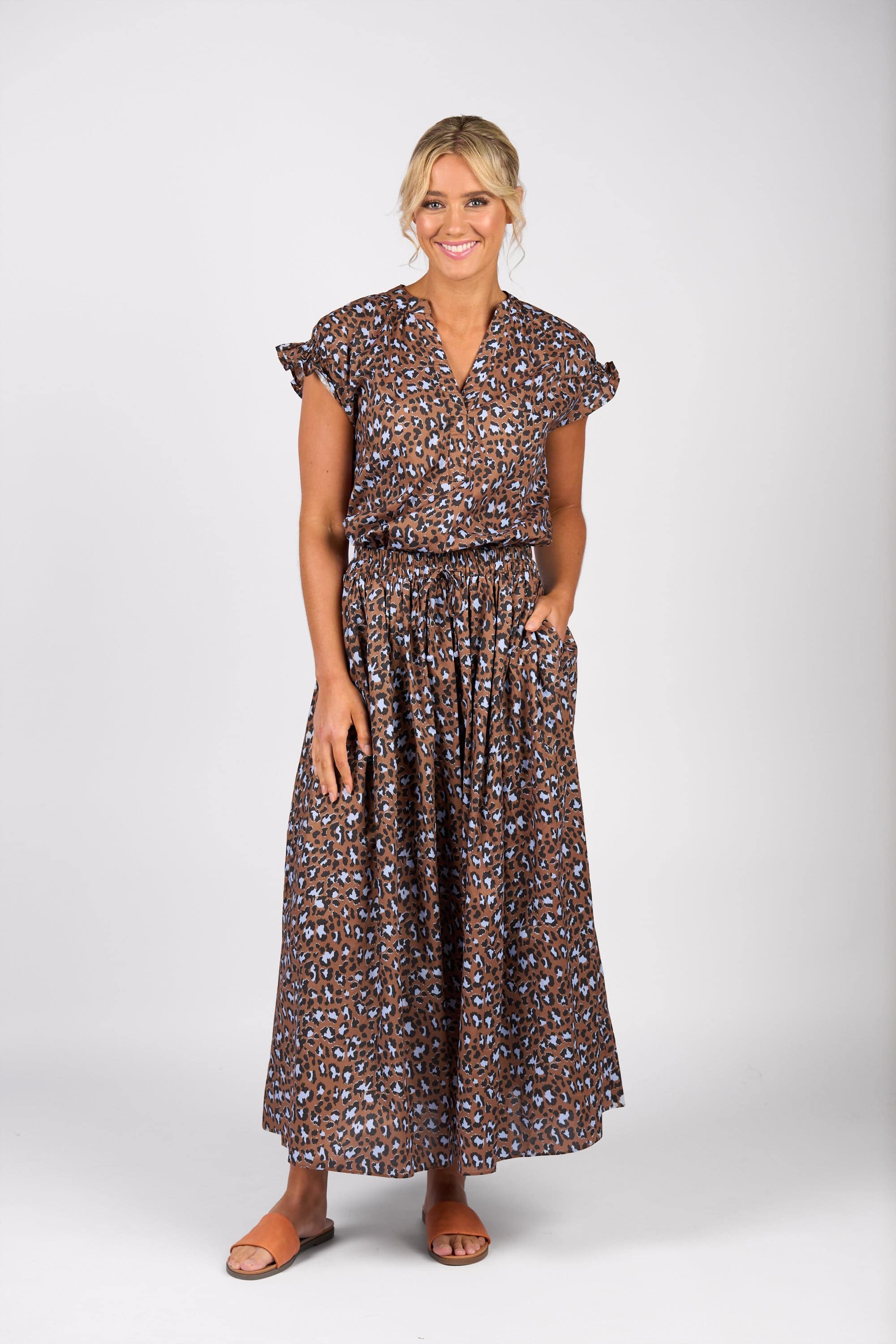 A woman with her blonde hair in a loose, low updo smiles against a white background. She wears the Vassalli Maxi Skirt - Nala in brown and blue leopard print with an elastic waistband, styled with short ruffled sleeves and tan sandals.