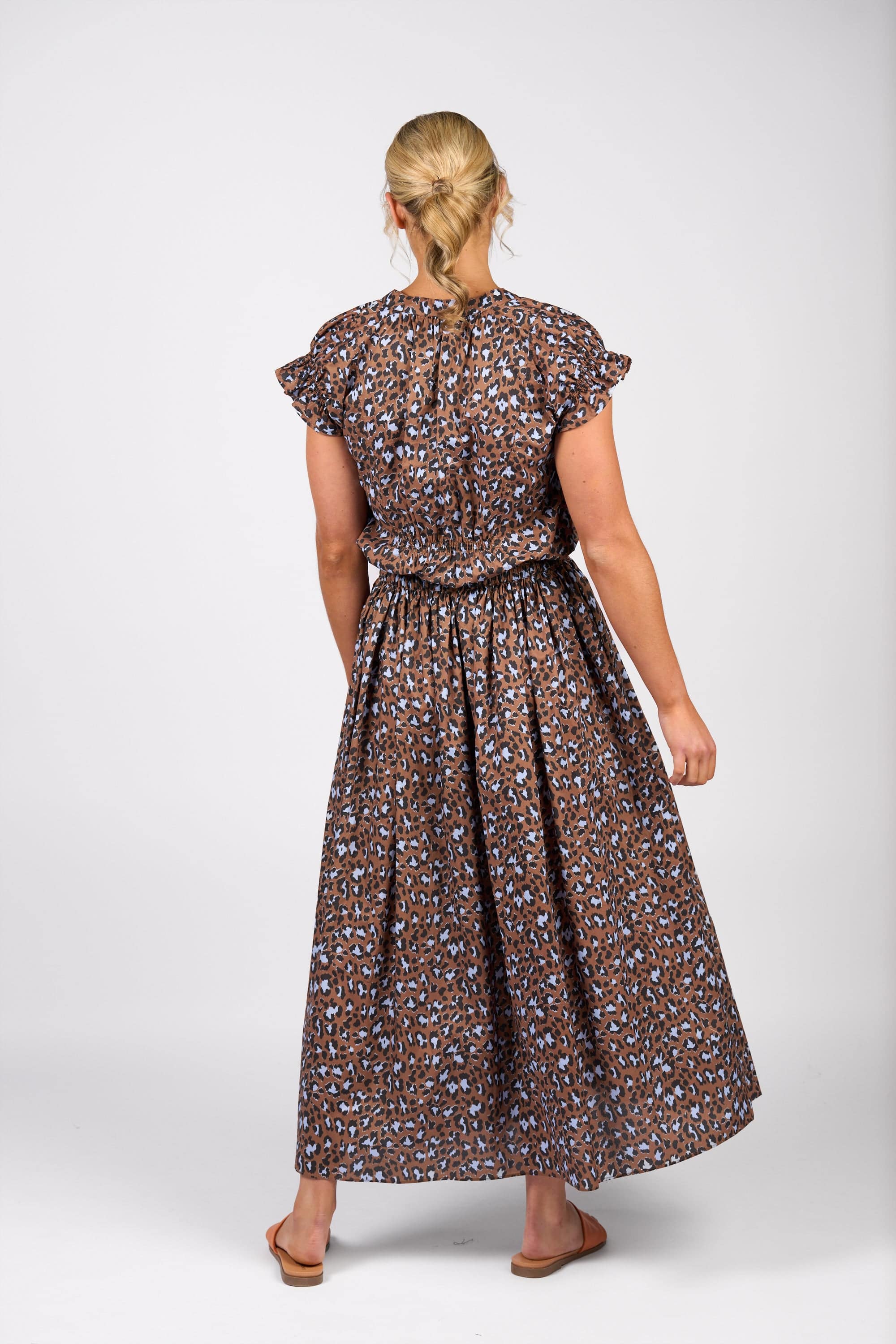 A woman faces away, wearing the Vassalli Maxi Skirt - Nala in a brown, blue, and black print. The skirt is cotton with an elastic waistband and a flowing silhouette. She pairs it with flat sandals, her blonde hair pulled back against a plain white background.