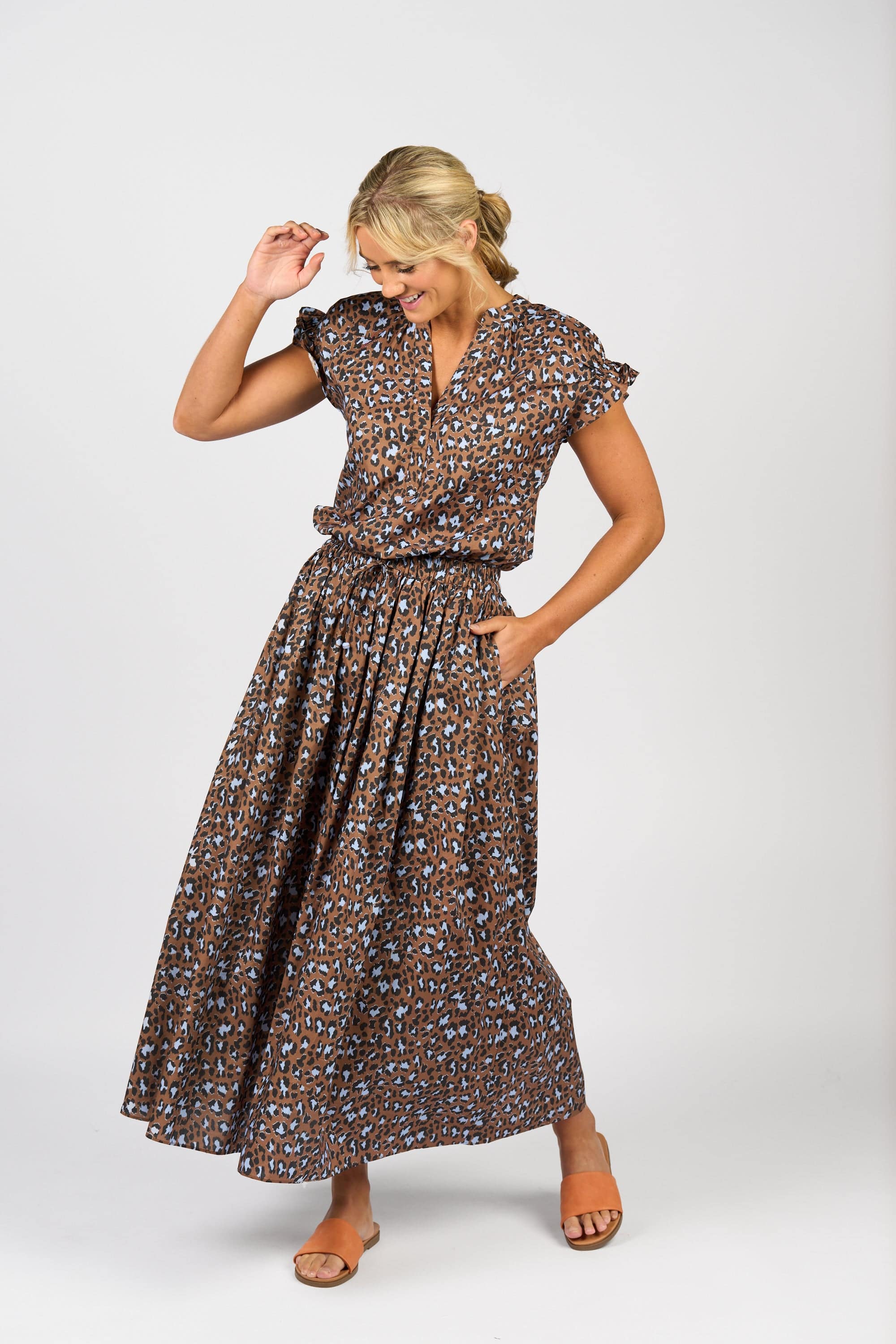 A woman in the Vassalli Maxi Skirt - Nala, featuring a brown, blue, and black animal print and elastic waistband, stands against a white background smiling, with one hand up and the other in her pocket. She completes the look with tan slide sandals.