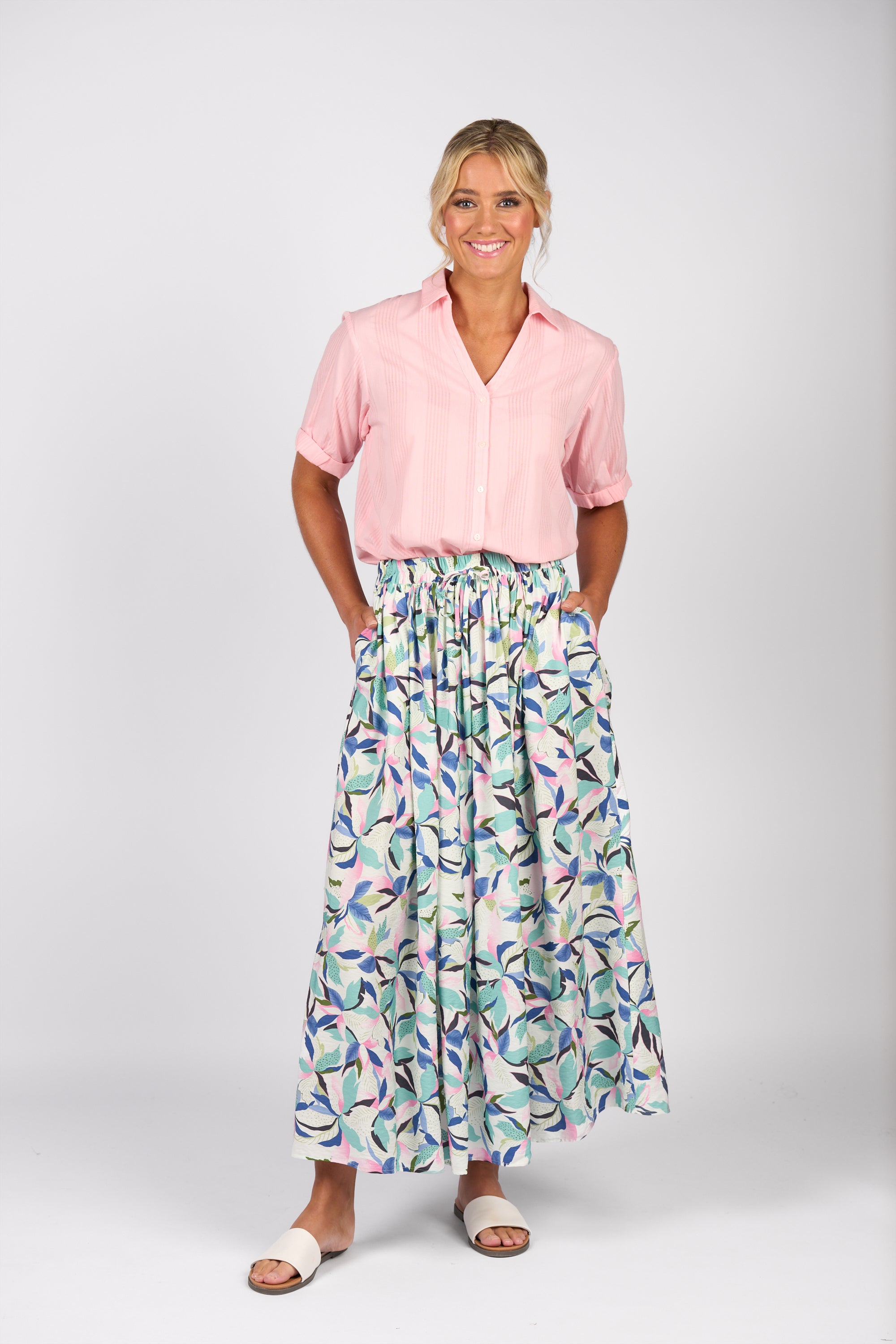 A smiling blonde woman wears the Vassalli Maxi Skirt - Soleil, featuring an elastic waistband and side seam pockets, paired with a light pink short-sleeve blouse and white sandals, standing against a plain white background.