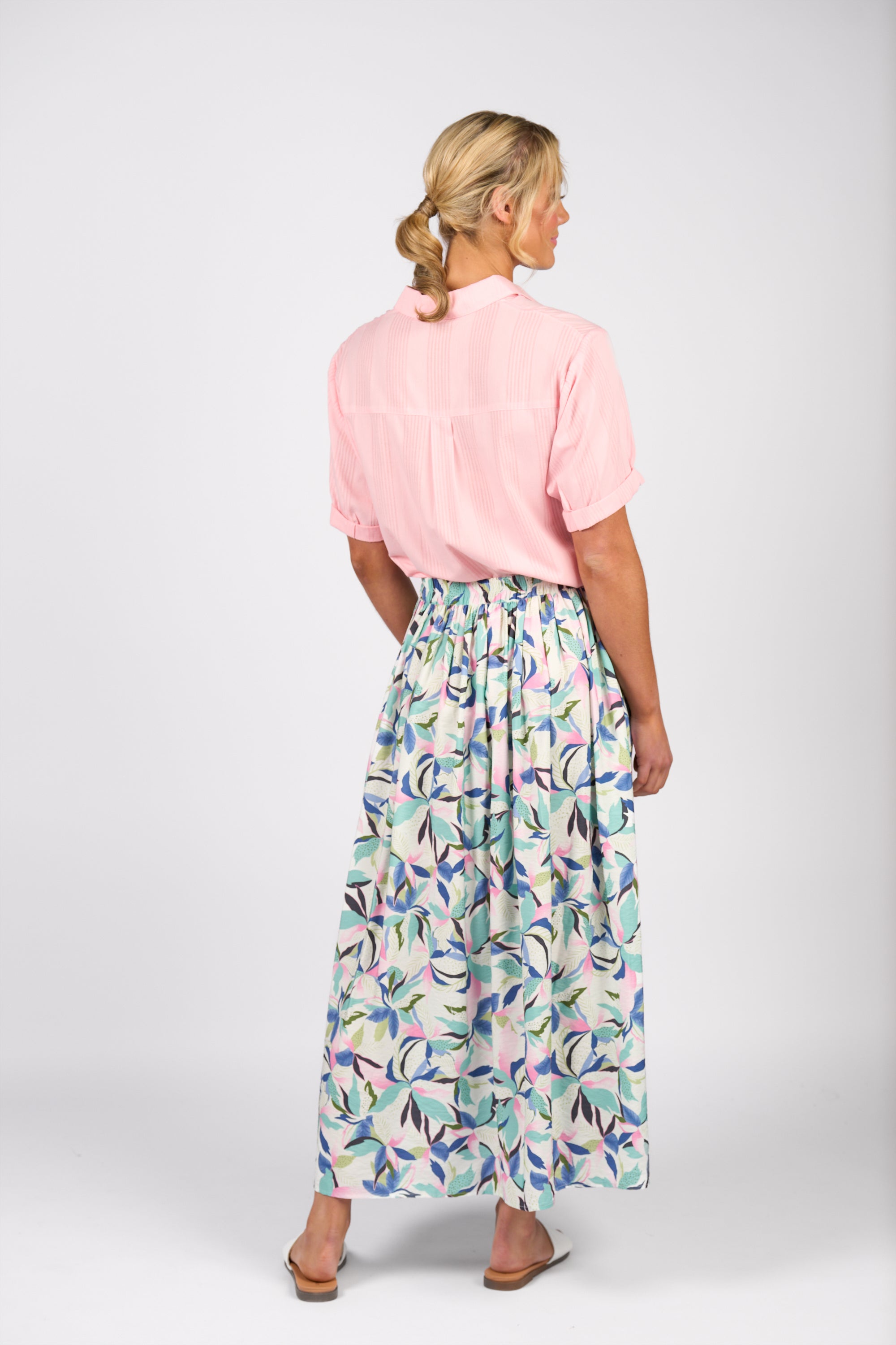 A woman with blonde hair tied back wears the Vassalli Maxi Skirt - Soleil, featuring a flowy print and side seam pockets, paired with a light pink short-sleeve blouse and sandals against a plain light background.