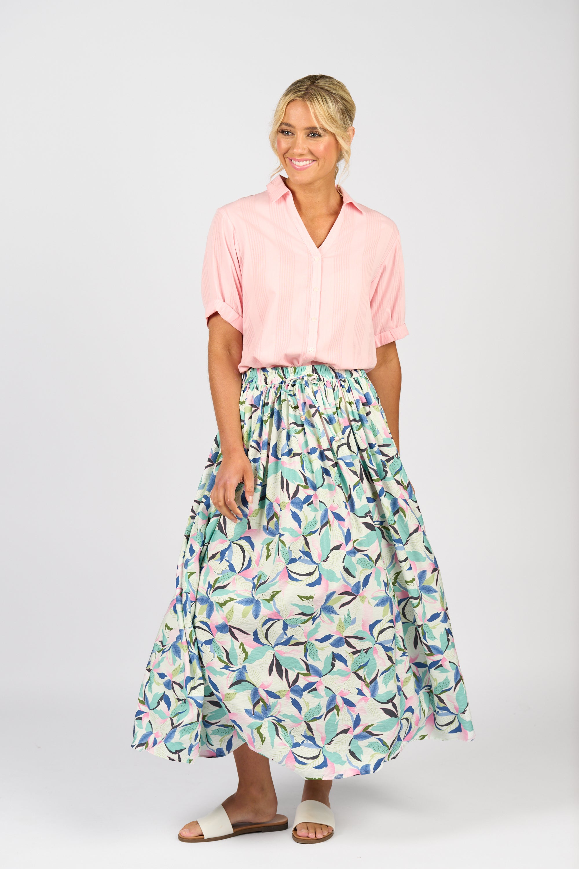 A woman smiles against a plain white background, wearing the Vassalli Maxi Skirt - Soleil with a colorful print and elastic waistband, paired with a light pink short-sleeve blouse and white sandals.