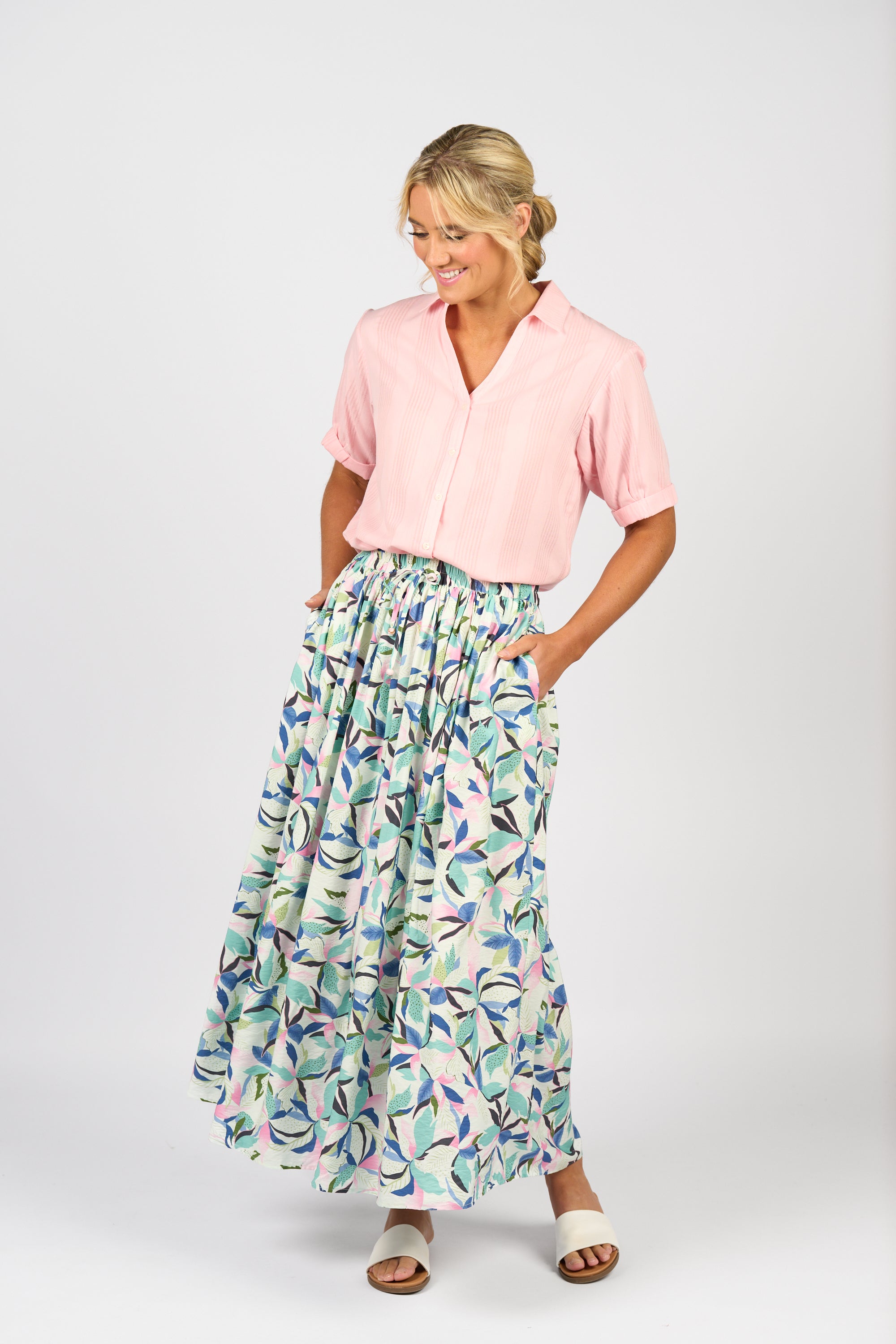 A woman stands against a plain background, smiling and looking down. She wears a light pink short-sleeve blouse, white sandals, and the Vassalli Maxi Skirt - Soleil, featuring an elastic waistband and side seam pockets.