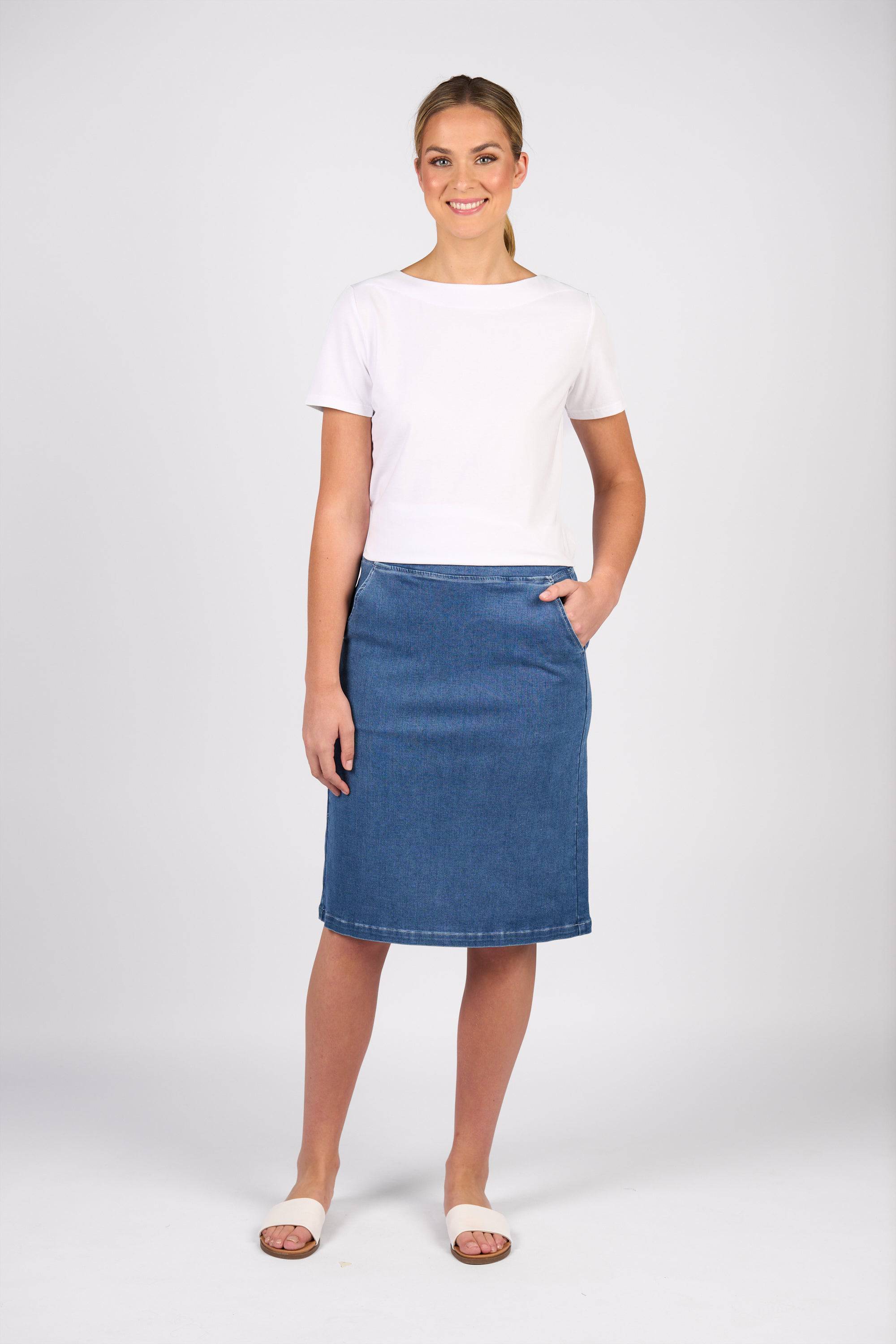 A woman stands smiling against a plain background, wearing the Vassalli Terra Skirt in Blue Hue Denim with a white short-sleeve top and open-toe sandals, one hand in her pocket, showcasing a relaxed casual style.