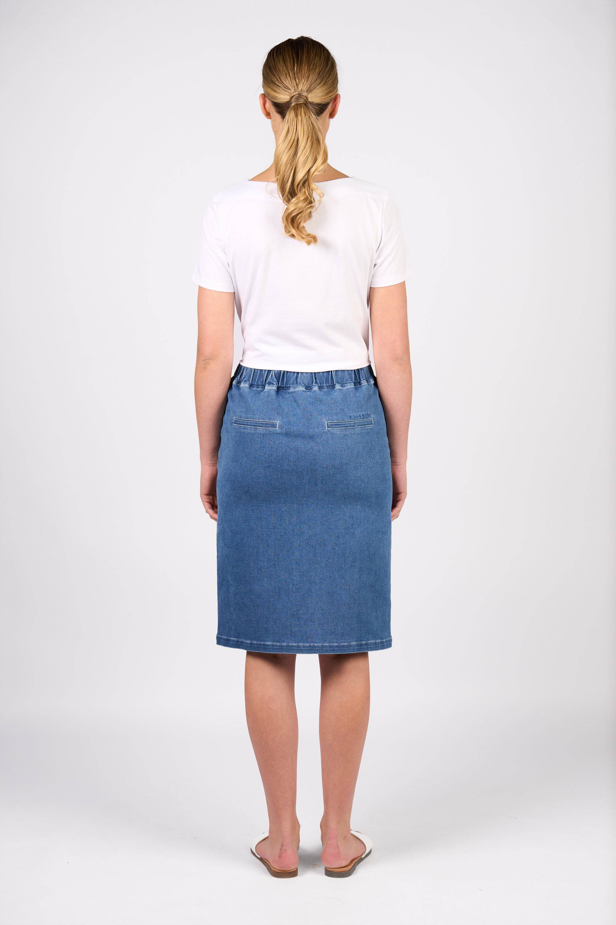 A woman with a low ponytail wears Vassalli's Terra Skirt in Blue Hue Denim, a knee-length stretch skirt with an elastic waistband, paired with a white short-sleeve top and white flats. She stands against a plain white background.