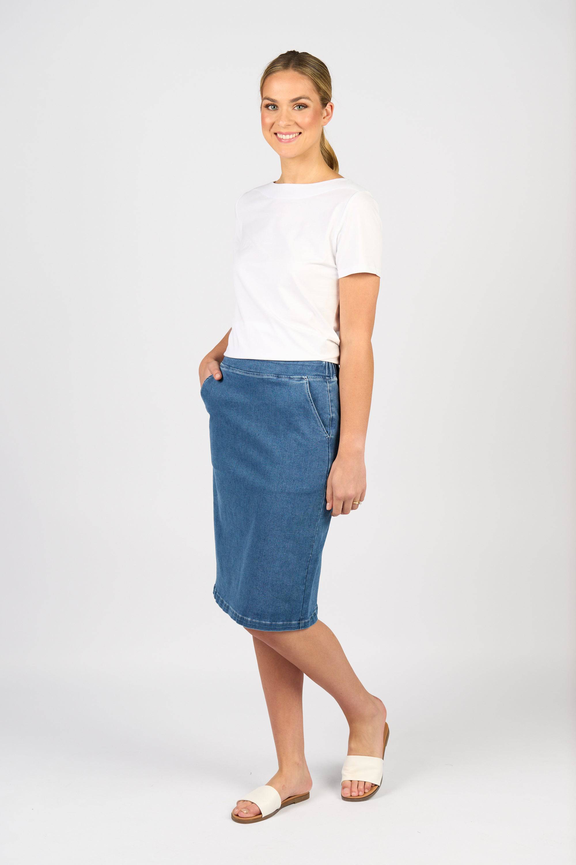 A woman smiles in a casual pose, wearing a white short-sleeve tee, the Vassalli Terra Skirt in Blue Hue Denim, white slide sandals, and her hair tied back in a ponytail against a plain light background.