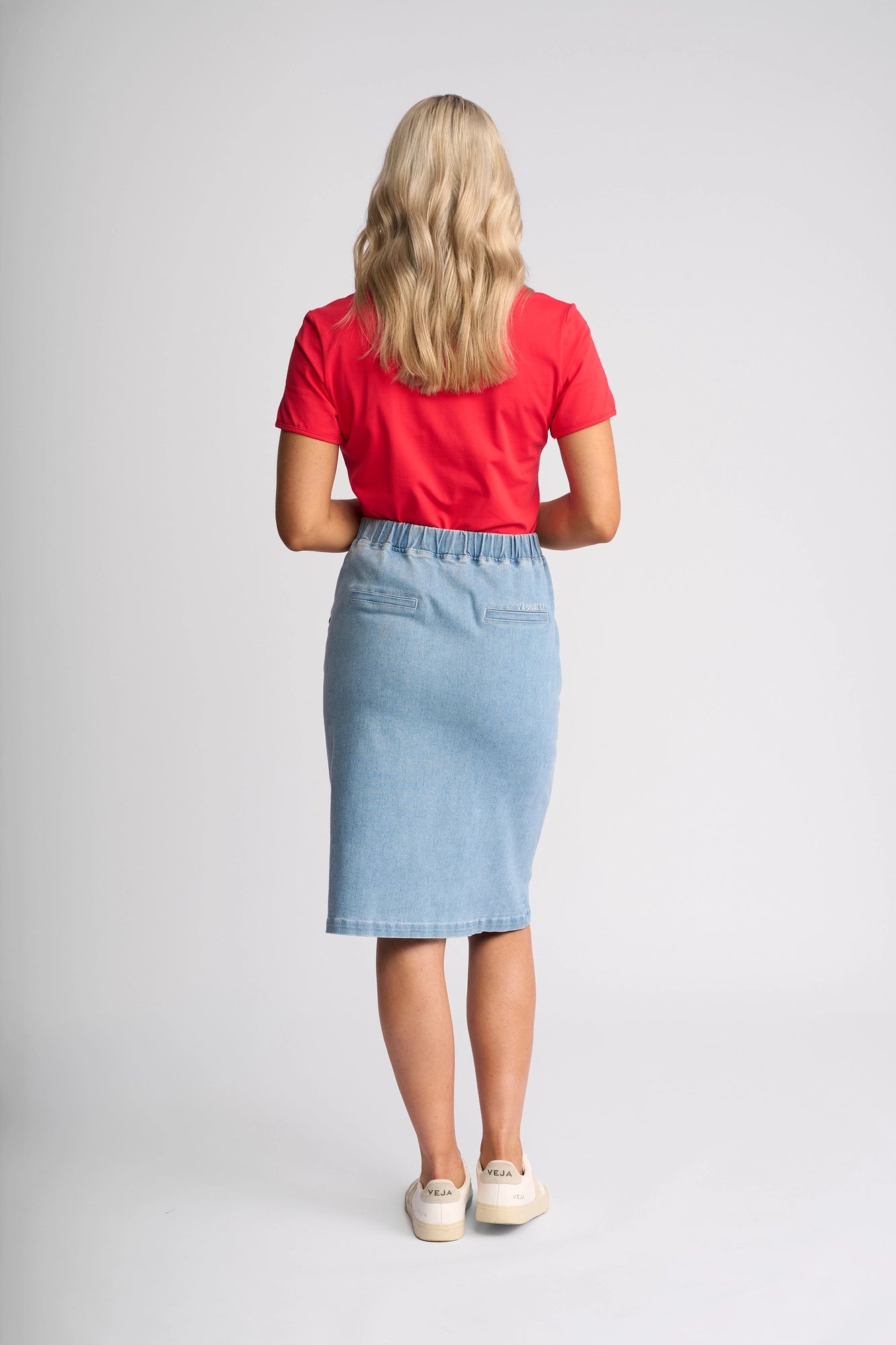 A woman with long blonde hair, wearing a red t-shirt, the Vassalli Terra Skirt in Light Wash Denim, and white slip-on shoes stands facing away from the camera against a plain white background.