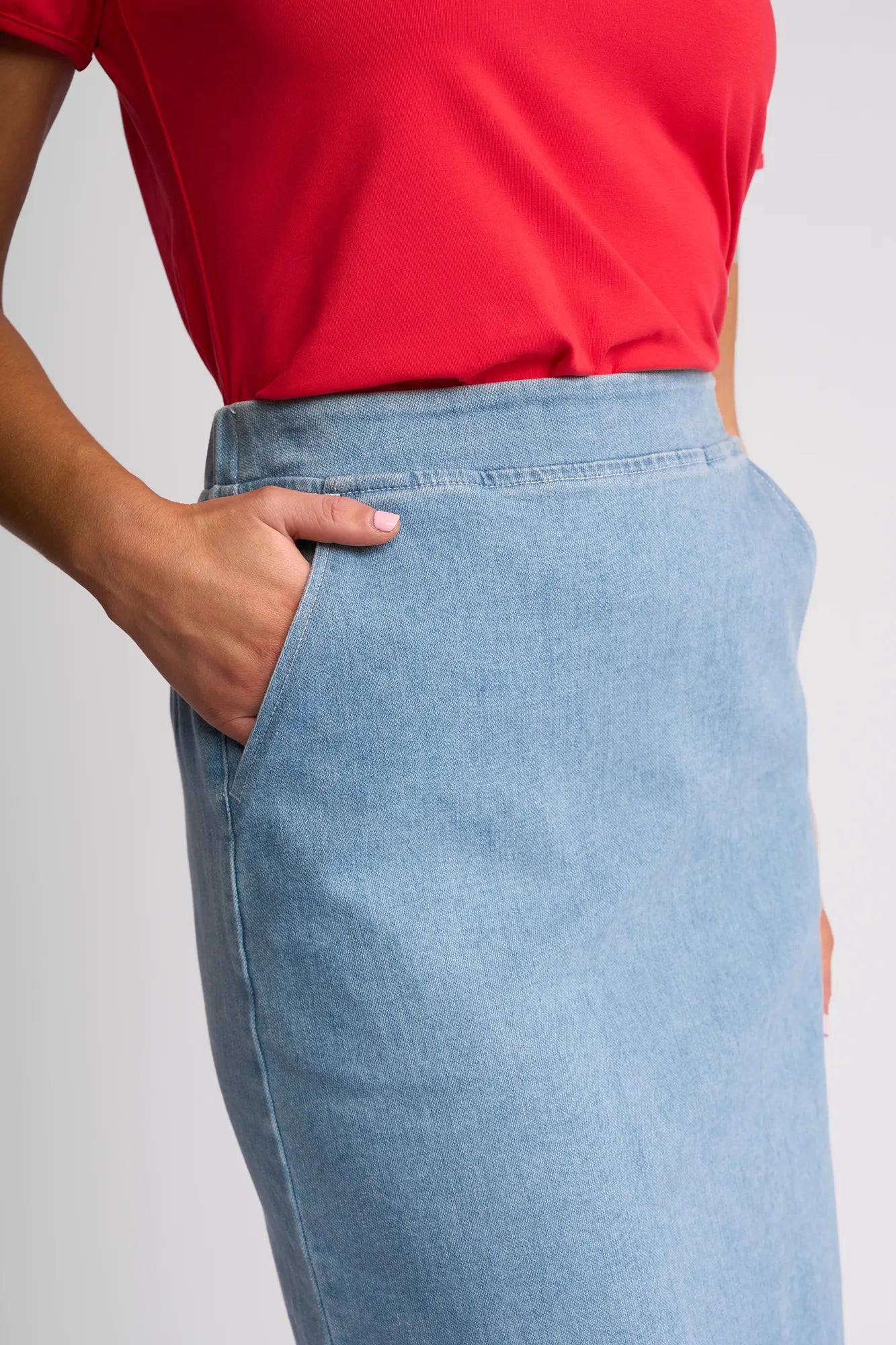 A person wearing a red top and the Vassalli Terra Skirt in light wash denim stands with one hand in their skirt pocket.