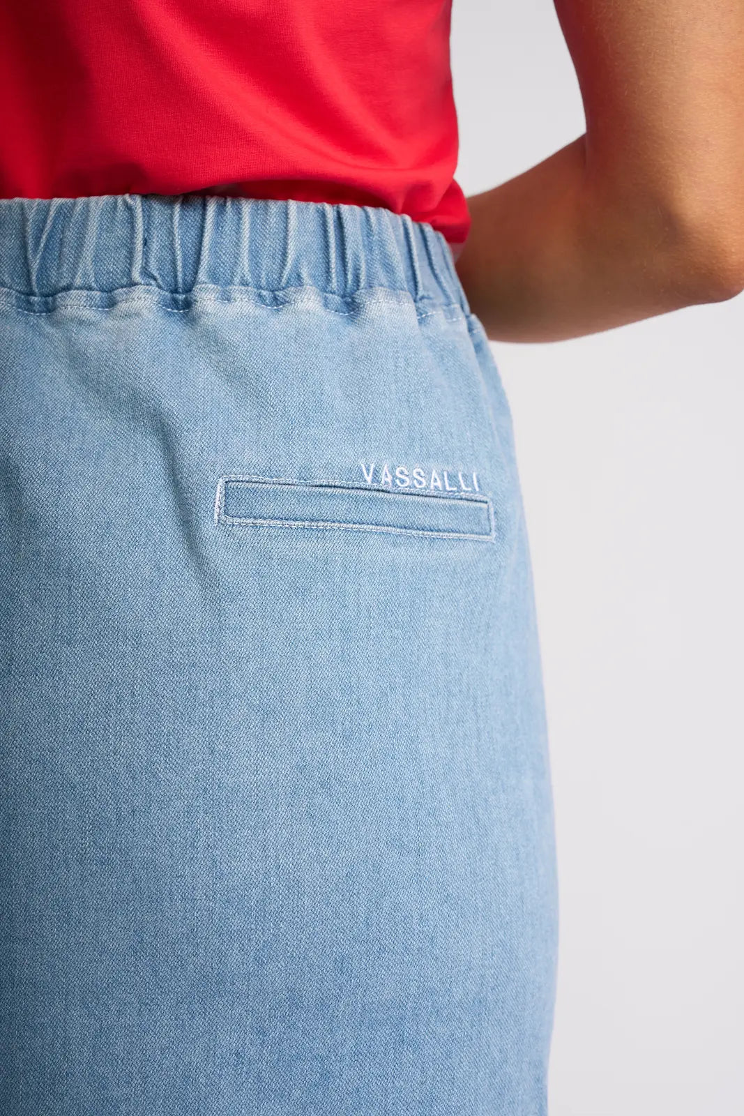 Close-up of a person wearing the Vassalli Terra Skirt in light wash denim, featuring an elastic waistband and a small back pocket with “VASSALLI” embroidered above it. This pull-on stretch denim skirt is paired with a red top.