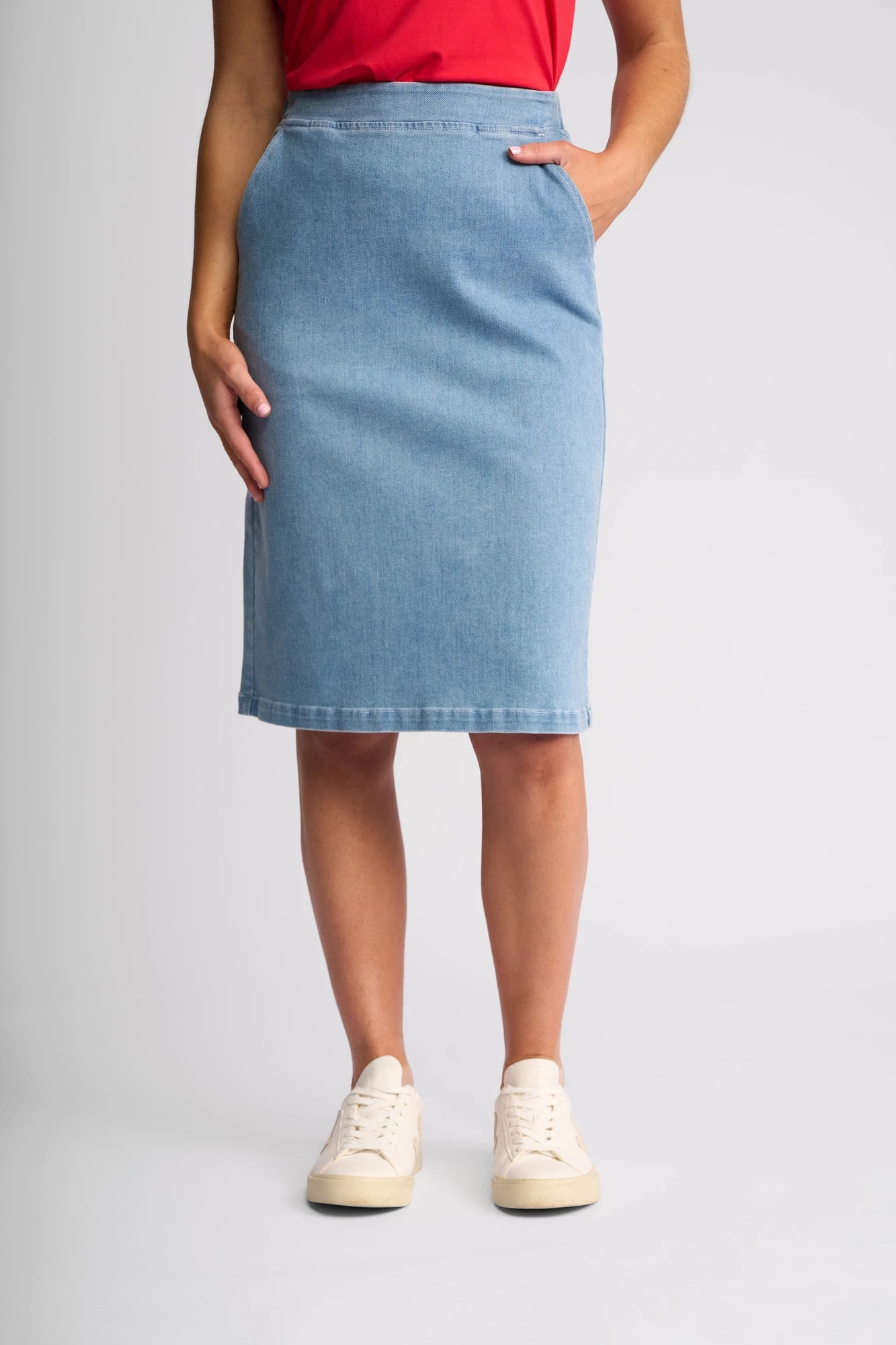 A person wearing a red top, the Vassalli Terra Skirt in Light Wash Denim, and white sneakers stands with one hand in a skirt pocket against a plain light background.
