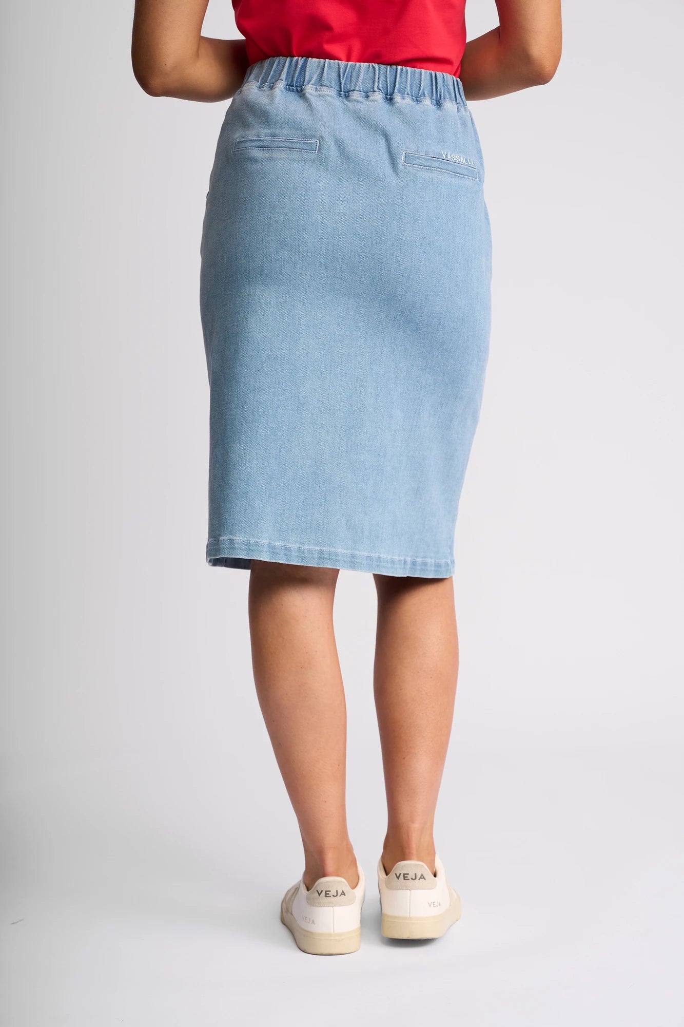 A person wearing a red top, the Vassalli Terra Skirt in Light Wash Denim that falls to the knee, and white Veja sneakers is shown from the back, standing against a plain light background.