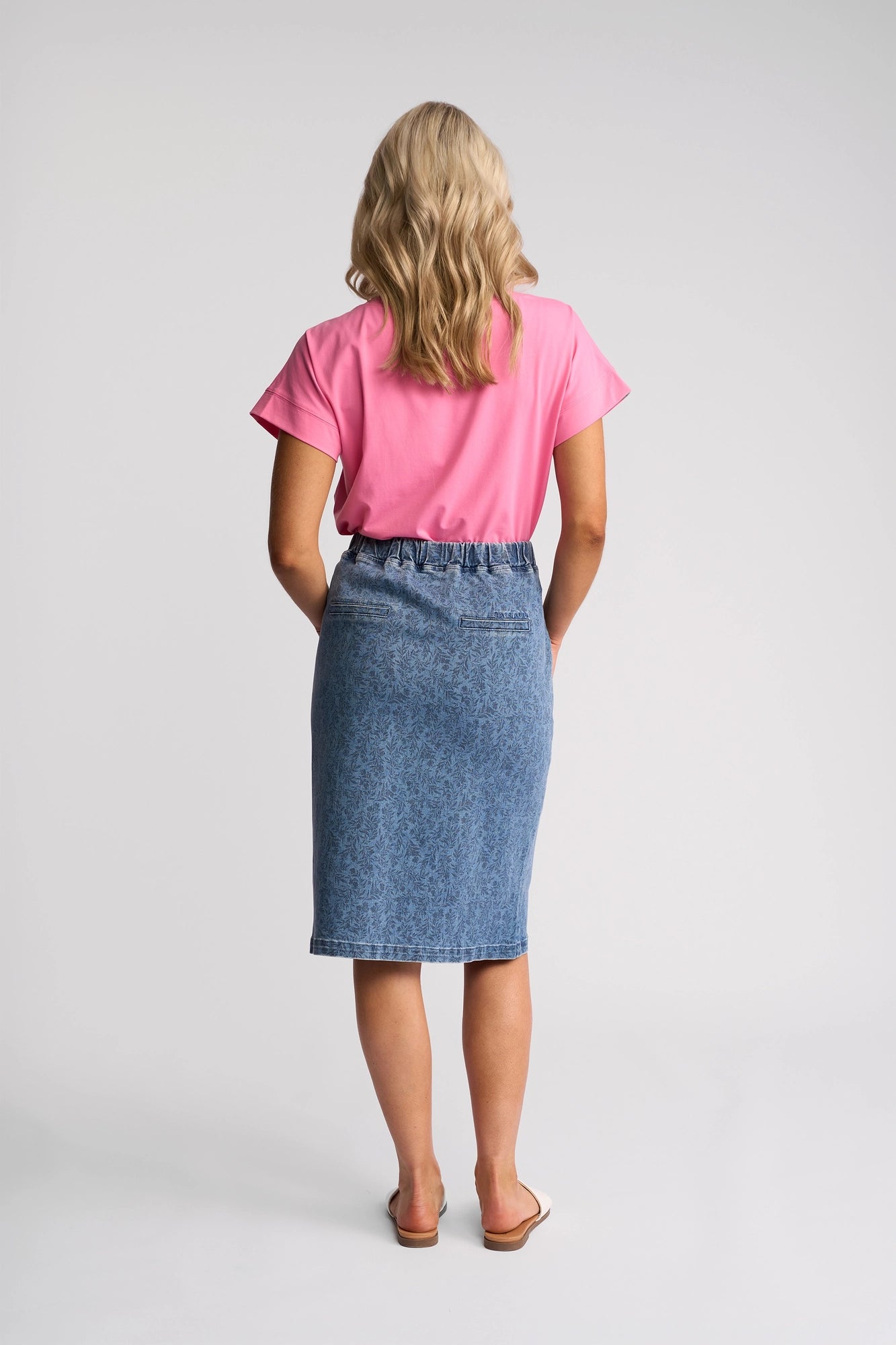 A woman stands facing away, wearing a pink short-sleeve shirt and the Vassalli Terra Skirt in Wisteria Denim—a stylish, knee-length stretch denim skirt with an elastic waistband. She completes her look with sandals, and her wavy blonde hair falls past her shoulders against a plain, light-colored background.