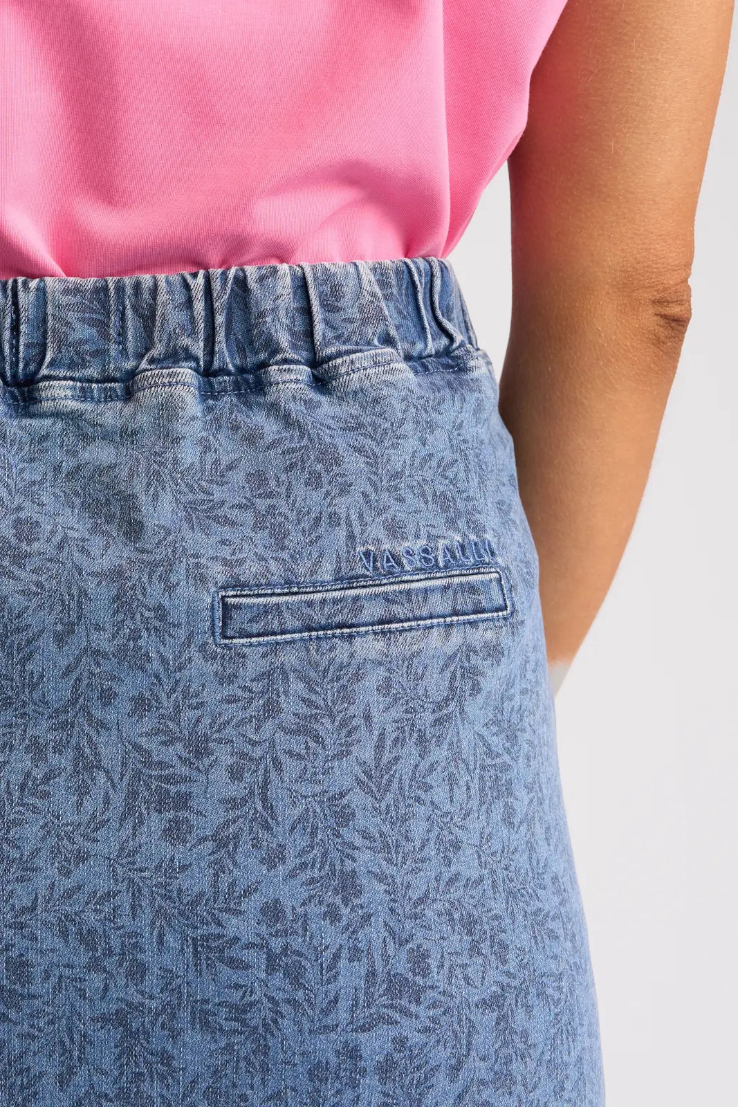 Close-up of a person wearing a pink shirt and the Vassalli Terra Skirt - Wisteria Denim, crafted from stretch denim with an elastic waistband and a small back pocket embroidered with “VASSALLI.” This skirt features a subtle floral print, adding a touch of elegance to the stylish look.