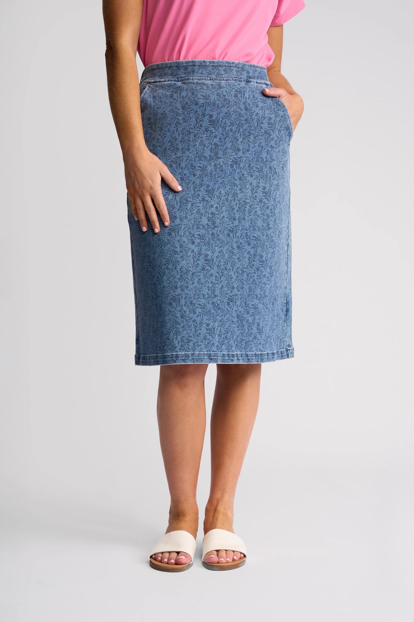 A person wearing a pink top, the Vassalli Terra Skirt in Wisteria Denim, and white slide sandals stands with one hand in a pocket against a plain light-colored background.