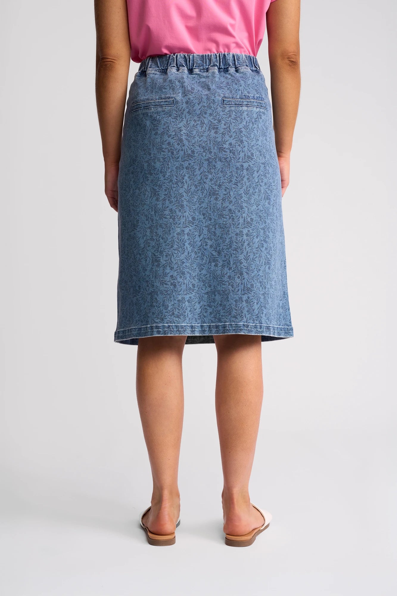 A person wearing a pink shirt, a blue Vassalli Terra Skirt - Wisteria Denim, and white slide sandals is standing facing away, showcasing the back of the outfit against a plain background.