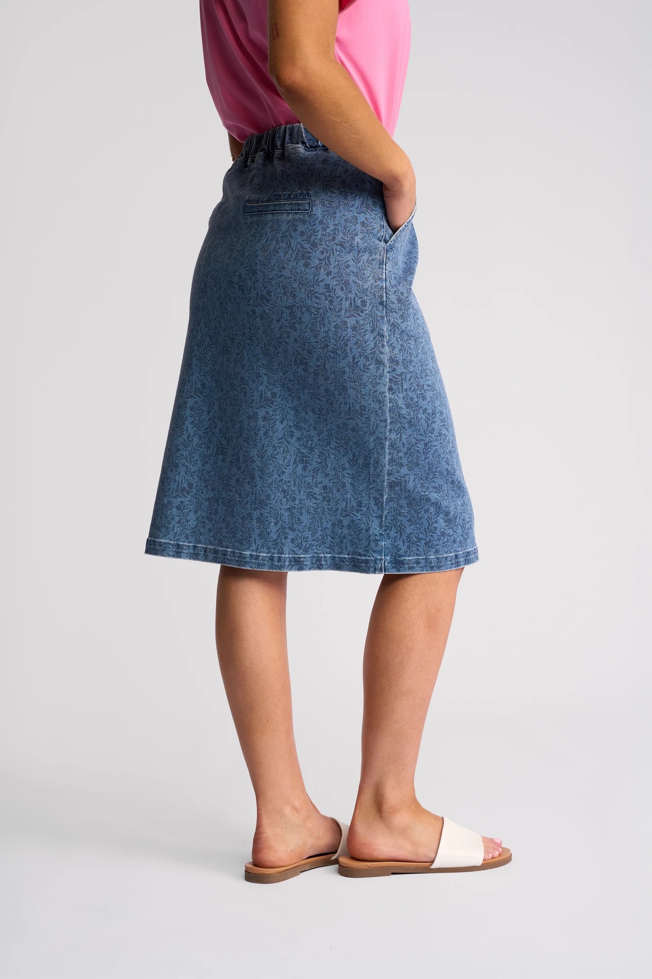 A person wearing a pink top, the Vassalli Terra Skirt - Wisteria Denim featuring a blue knee-length stretch denim with a subtle floral print, and white slide sandals, stands with one hand in a pocket, viewed from the side against a plain background.