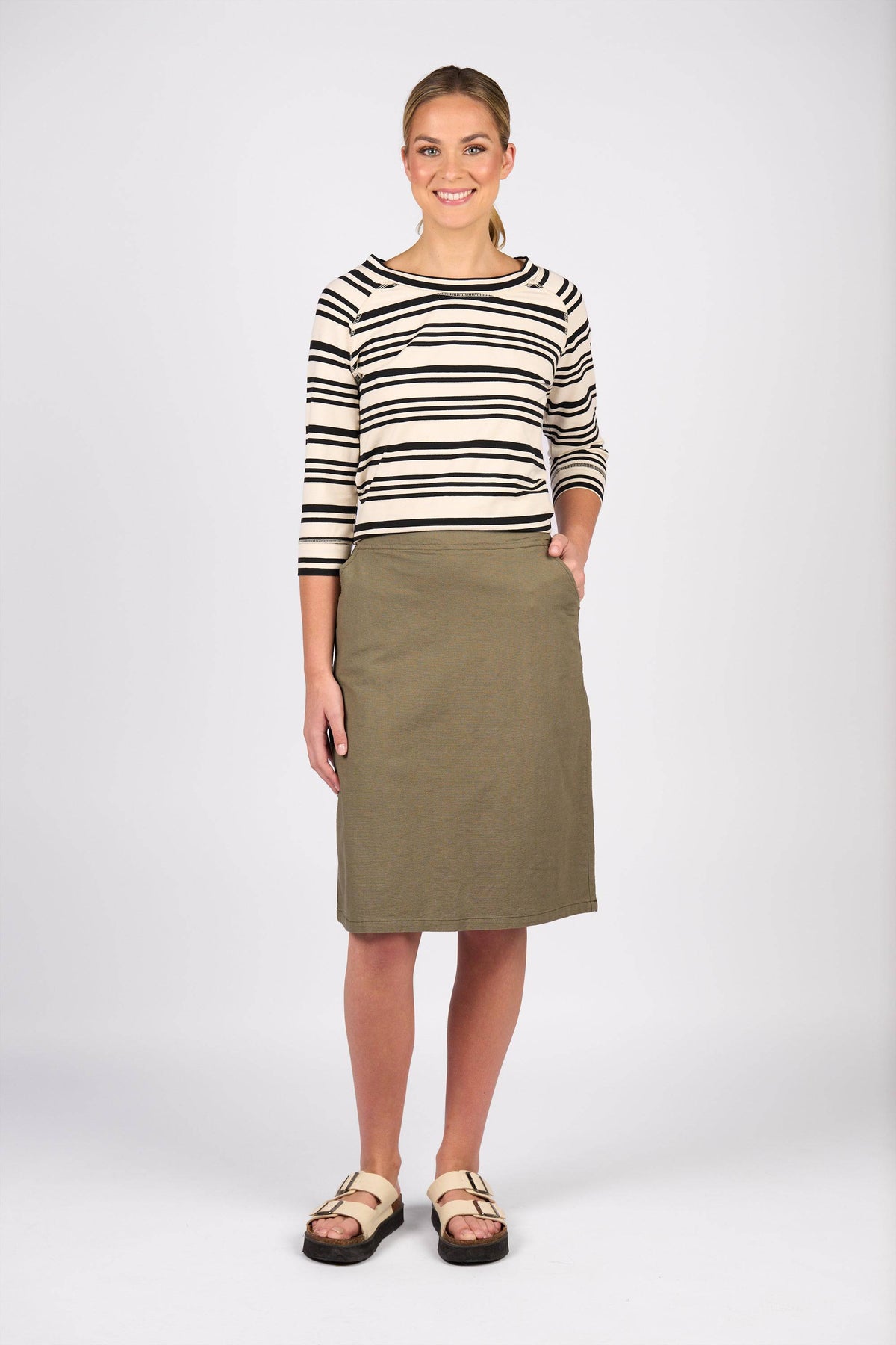 A woman smiles, wearing a black and white striped long-sleeve shirt, the Vassalli Belen Skirt - Army, and beige sandals. One hand is in her skirt pocket as she stands against a plain white background.
