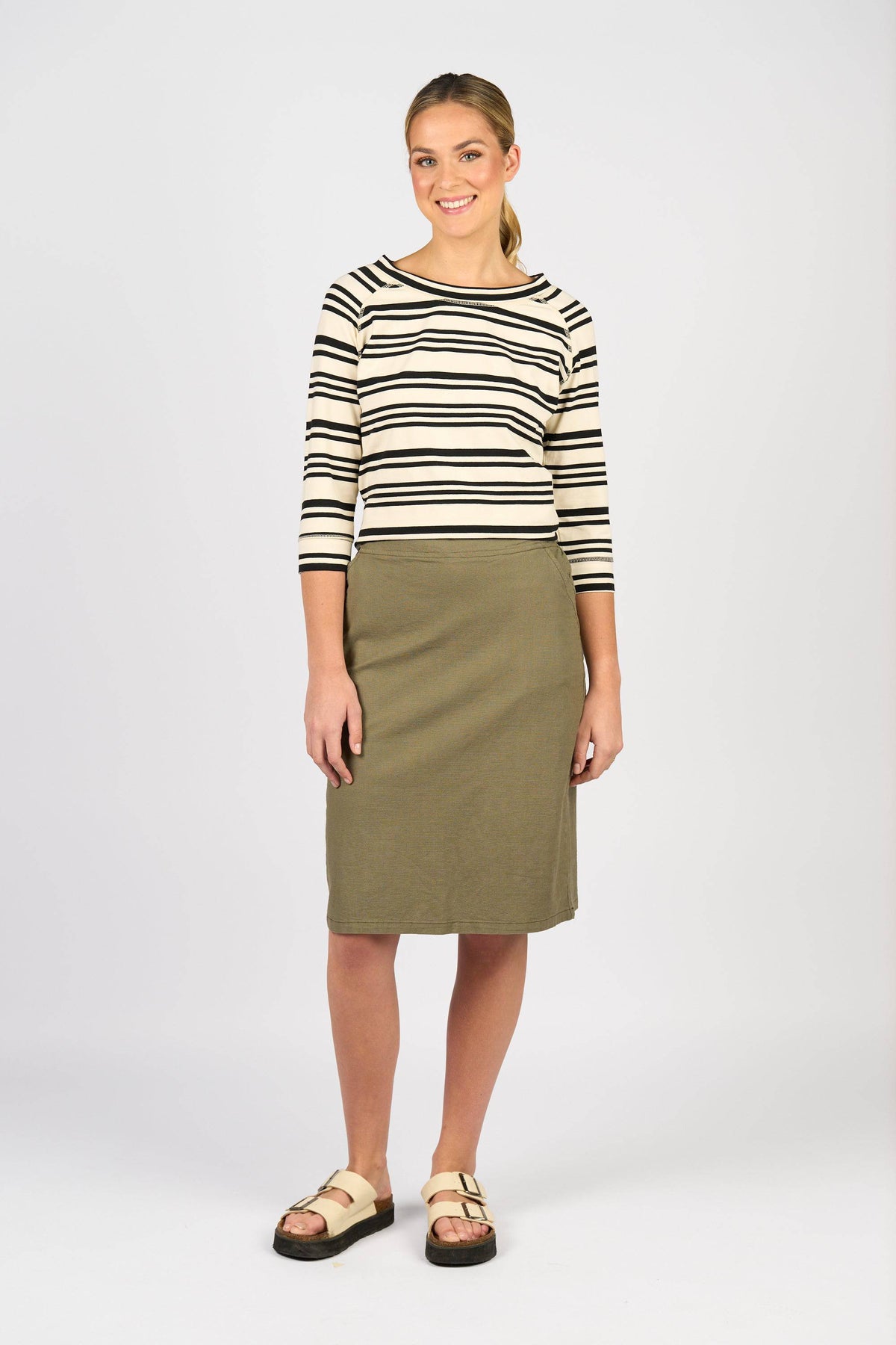A woman smiles against a light grey background, wearing a cream and black striped 3/4 sleeve top, the Vassalli Belen Skirt - Army, and beige buckle sandals.