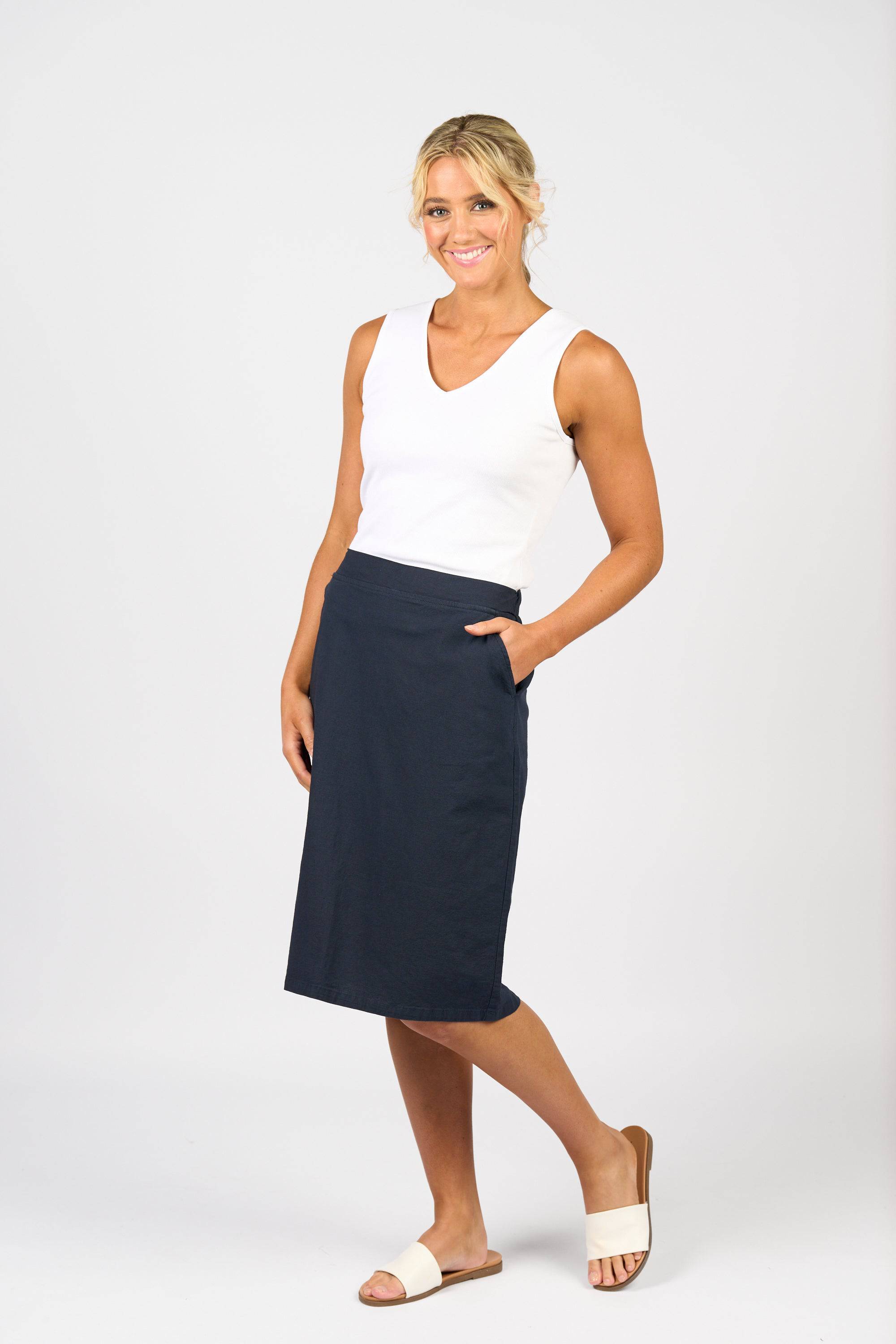 A woman smiles, wearing the Vassalli Belen Skirt - Ink with an elastic waistband and pockets, paired with a white sleeveless top and beige slide sandals. Her blonde hair is styled up and she stands against a plain white background.