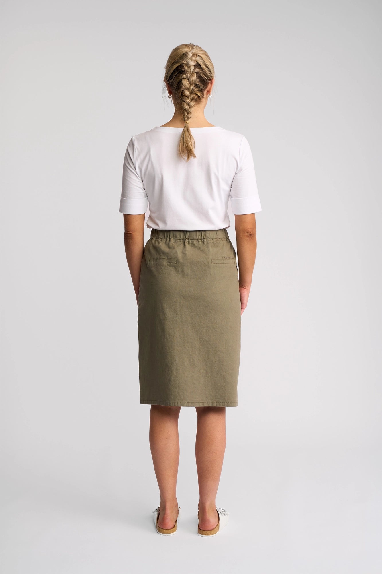 A woman faces away, her blonde hair styled in two braids, wearing a white short-sleeve shirt, sandals, and the Vassalli Belen Skirt - Army. Crafted from stretch cotton with a lightweight pull-on design, the skirt provides a comfortable and relaxed look against the plain light-colored background.