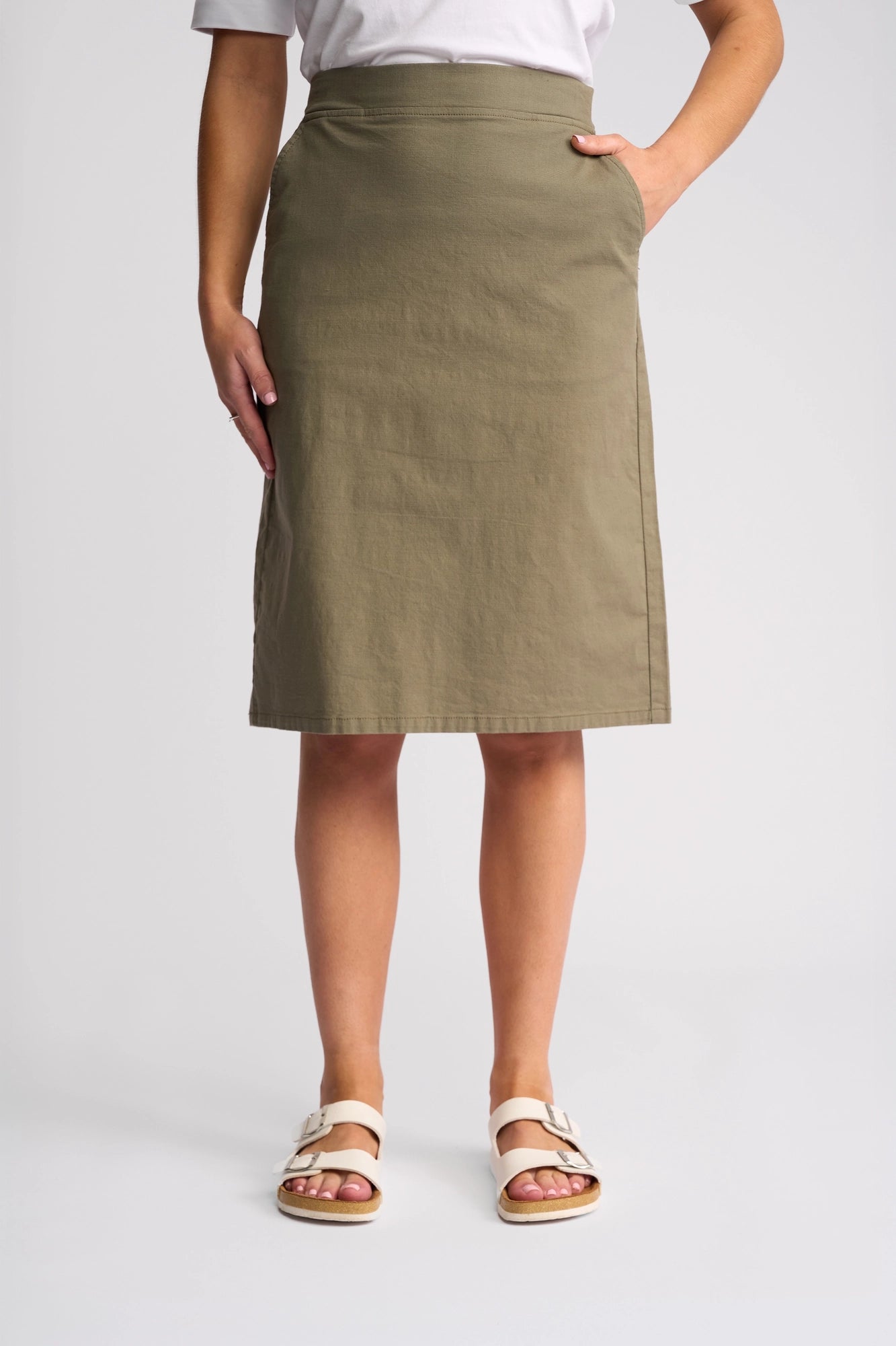 A person stands and poses in the Vassalli Belen Skirt - Army, paired with a white top and white sandals against a plain background. Their left hand is casually tucked into the skirt pocket, showcasing the ease and comfort of this lightweight pull-on skirt.