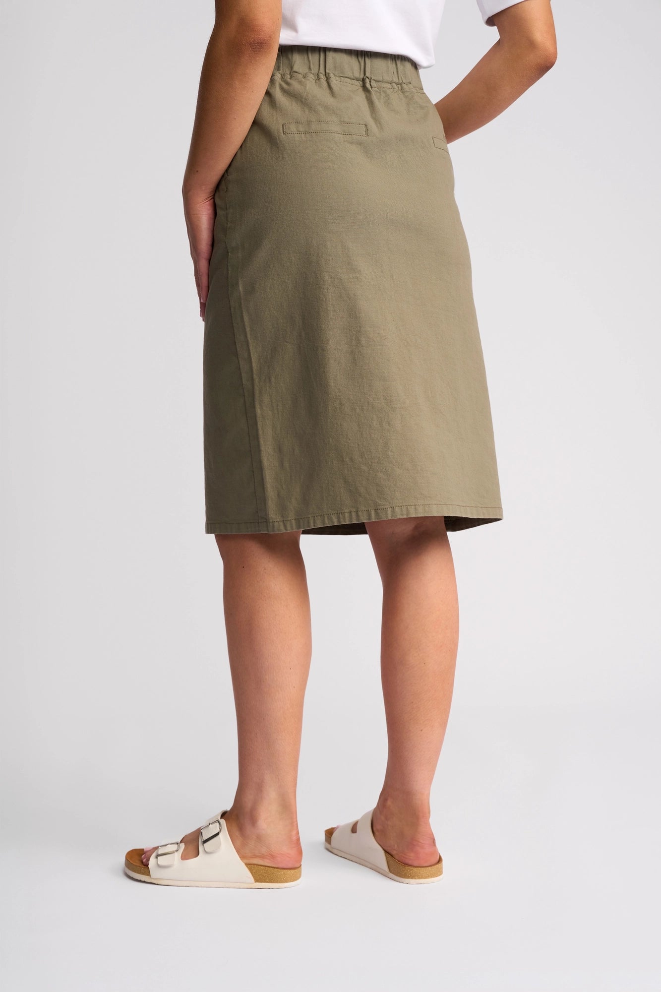 Person wearing a Vassalli Belen Skirt in Army—a lightweight pull-on, stretch cotton skirt—paired with a white T-shirt and white sandals, standing against a plain light background with hands in skirt pockets, shown from the back.