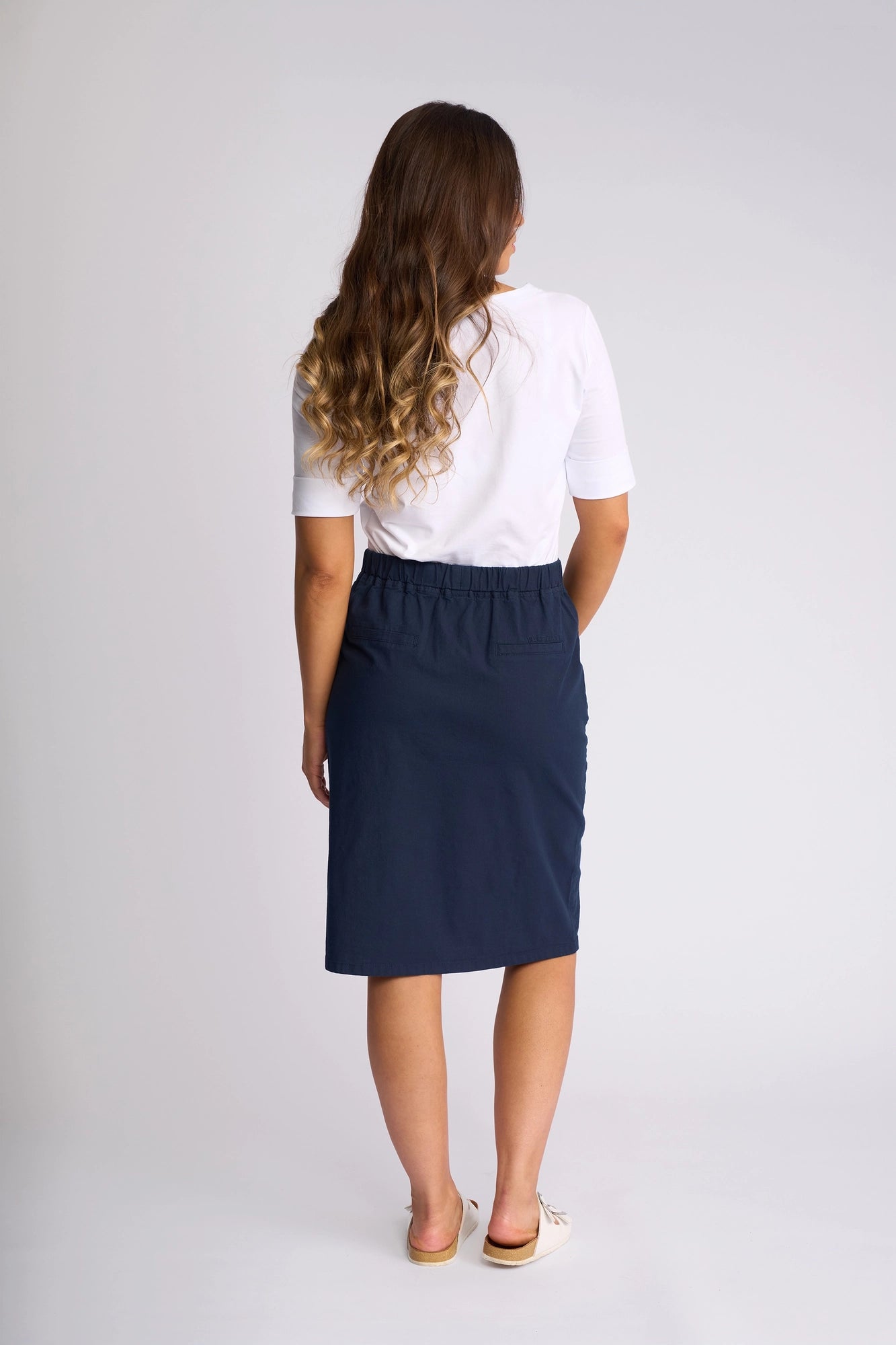 A woman with long wavy hair stands facing away, wearing a white short-sleeve shirt, the Vassalli Belen Skirt - Ink crafted from stretch cotton for comfort, and beige slip-on shoes against a plain white background.