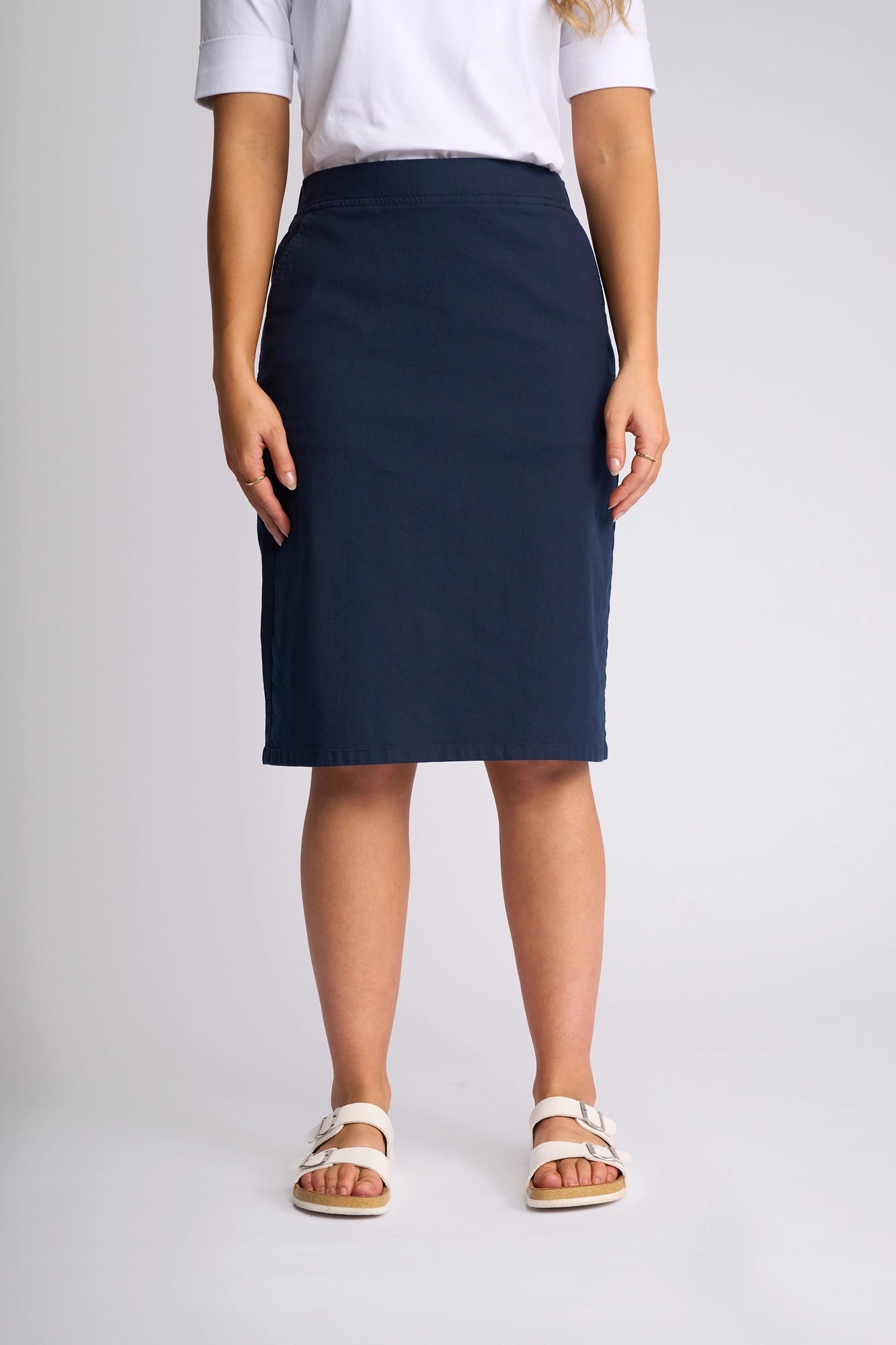 A woman stands facing the camera against a plain light background, wearing a white short-sleeve shirt, the Vassalli Belen Skirt - Ink crafted from stretch cotton, and white sandals.