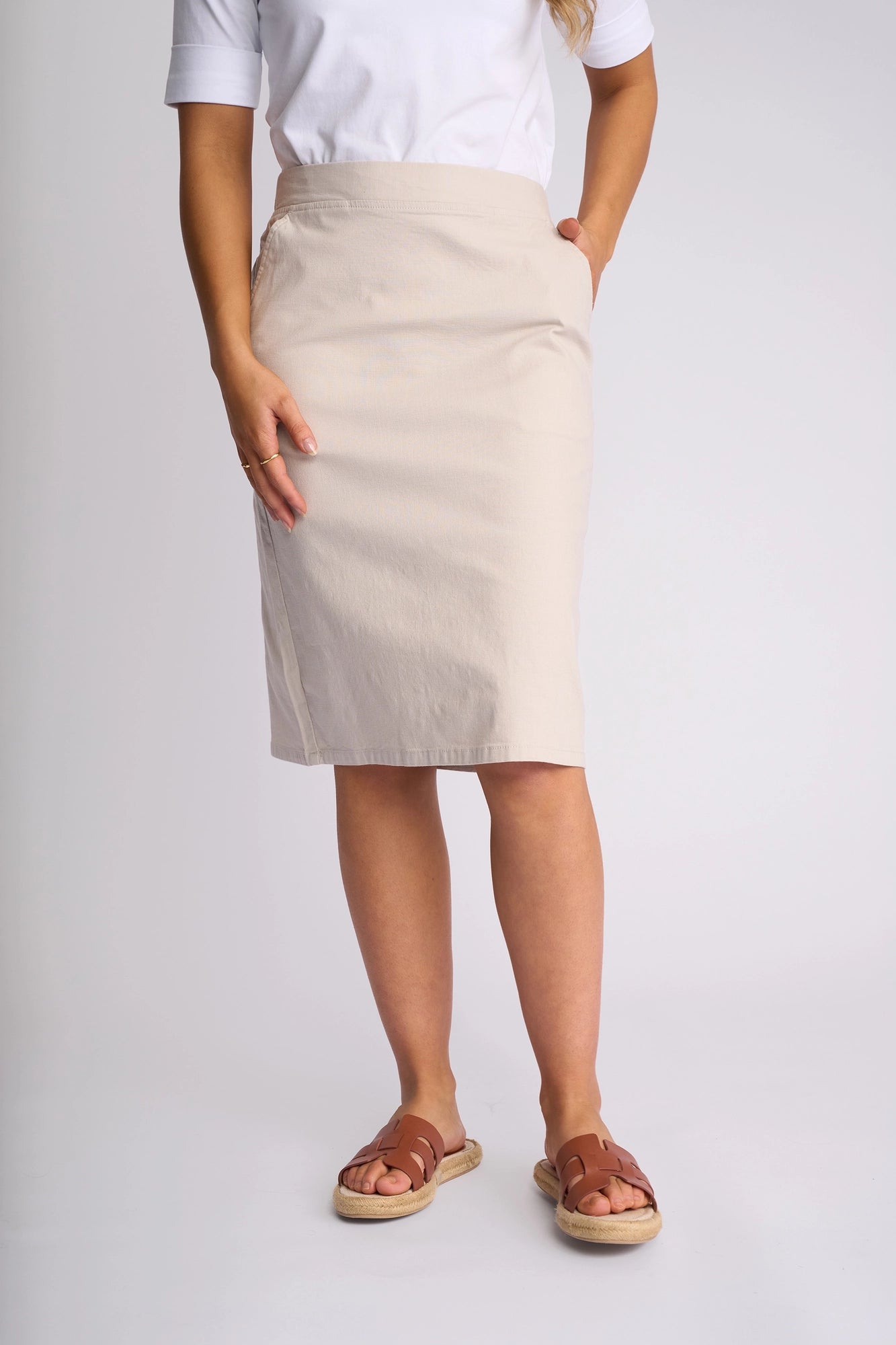 A woman stands with her hands in her pockets, wearing a white shirt, the Vassalli Belen Skirt in Stone—a knee-length beige skirt—and brown sandals. The background is plain and light-colored, and her head is not visible in the image.
