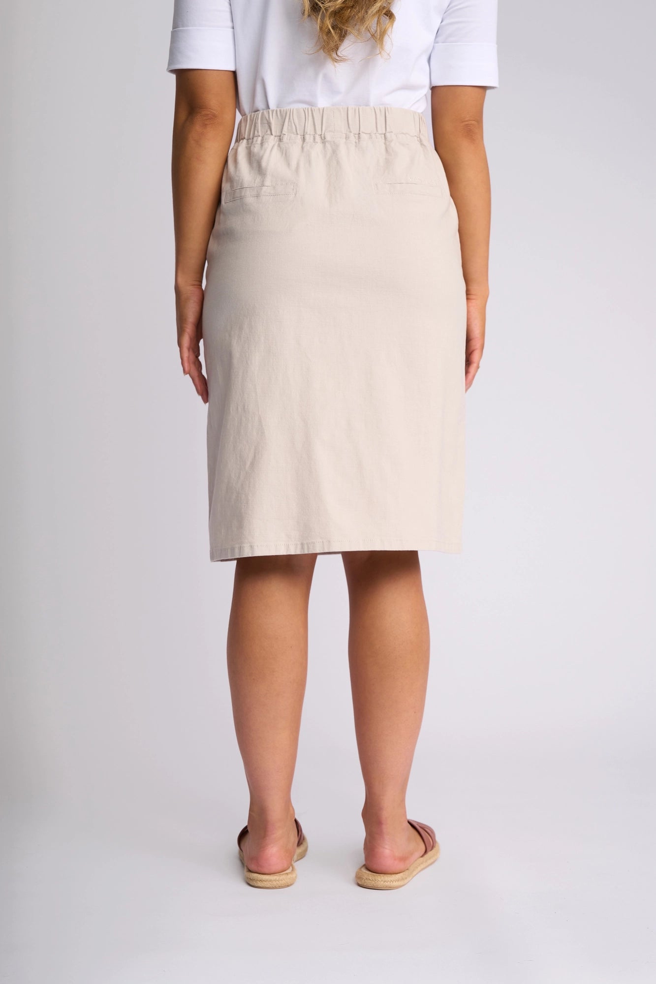 A woman stands facing away, wearing a white shirt and the Vassalli Belen Skirt in Stone—a beige, knee-length skirt with an elastic waistband—paired with tan slip-on shoes. Her wavy hair is partly visible, and the background is plain light gray.