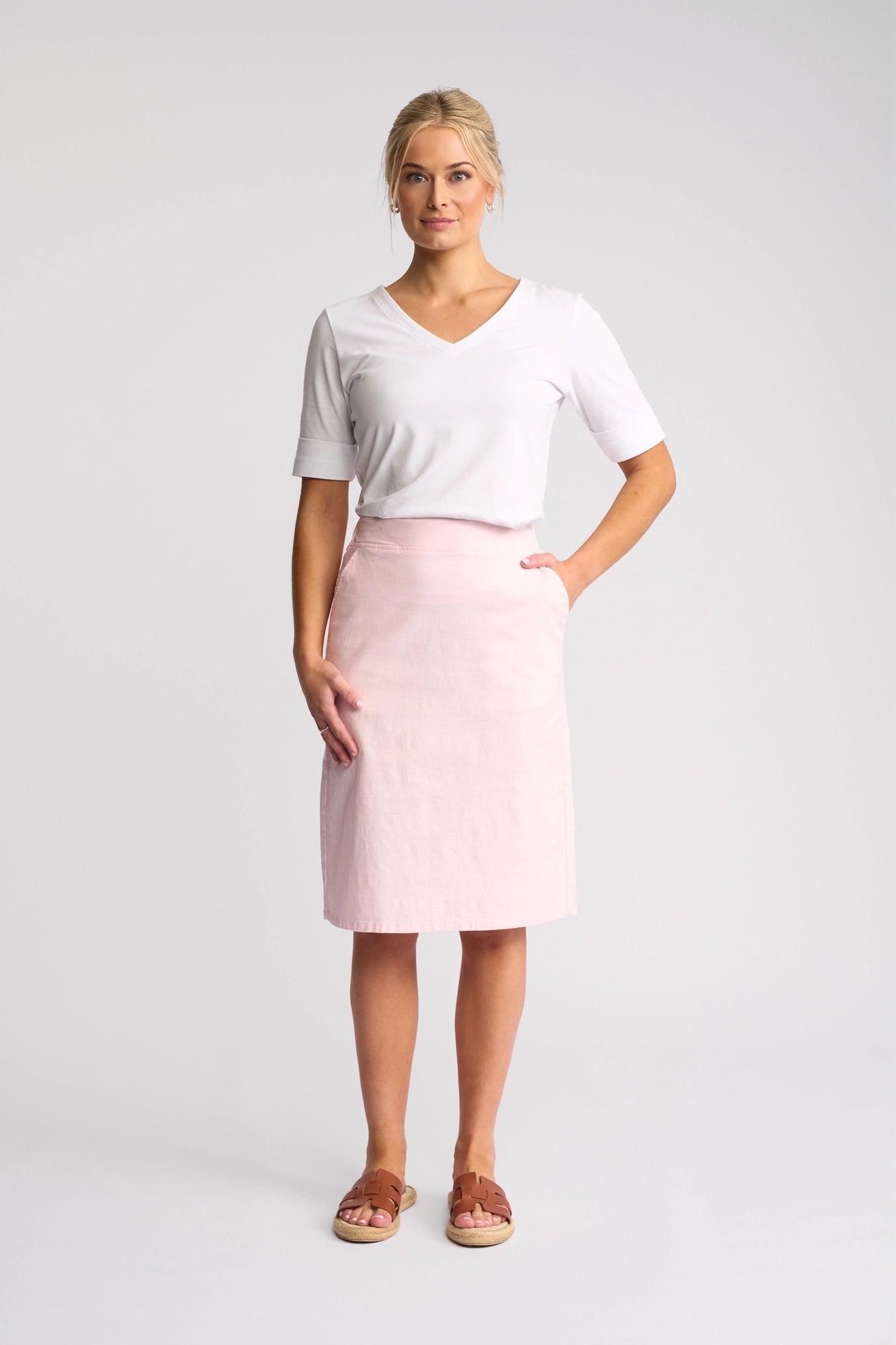 A woman stands against a plain background, wearing a white V-neck top and the Vassalli Belen Skirt in Taffy—a lightweight pull-on skirt made from stretch cotton. With her blonde hair tied back, she has one hand in her skirt pocket and looks straight ahead with a slight smile.
