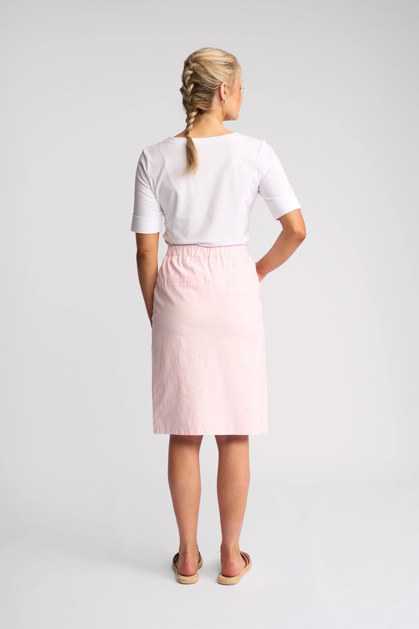 A woman with blonde hair in a braid, wearing a white top, the Vassalli Belen Skirt - Taffy—an elegant, lightweight pull-on skirt—and sandals stands facing away in a brightly lit studio with a plain white background.