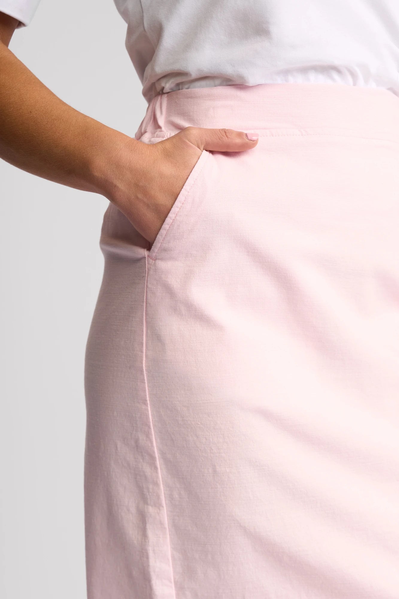 A person wearing a Vassalli Belen Skirt in Taffy and a white top has their right hand placed in the skirt pocket. The background is plain and light-colored.