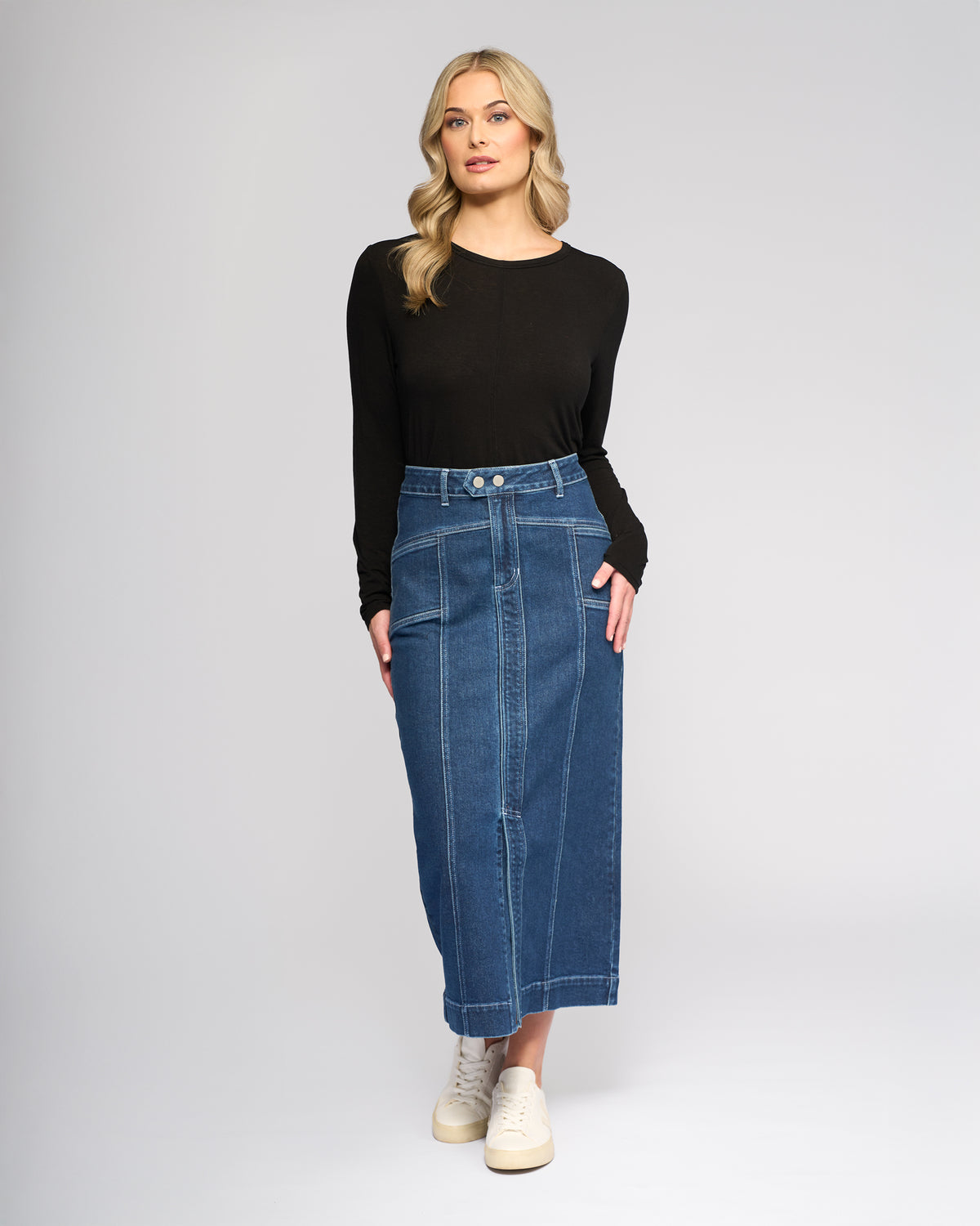 A woman with wavy blonde hair and a neutral expression wears a black long-sleeve top, white sneakers, and the Vassalli Rook Skirt in Dark Blue, featuring a panelled design, against a plain background.