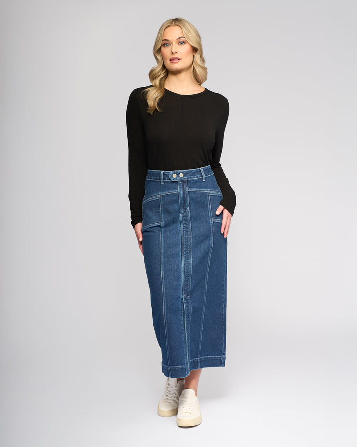 A woman with wavy blonde hair and a neutral expression wears a black long-sleeve top, white sneakers, and the Vassalli Rook Skirt in Dark Blue, featuring a panelled design, against a plain background.