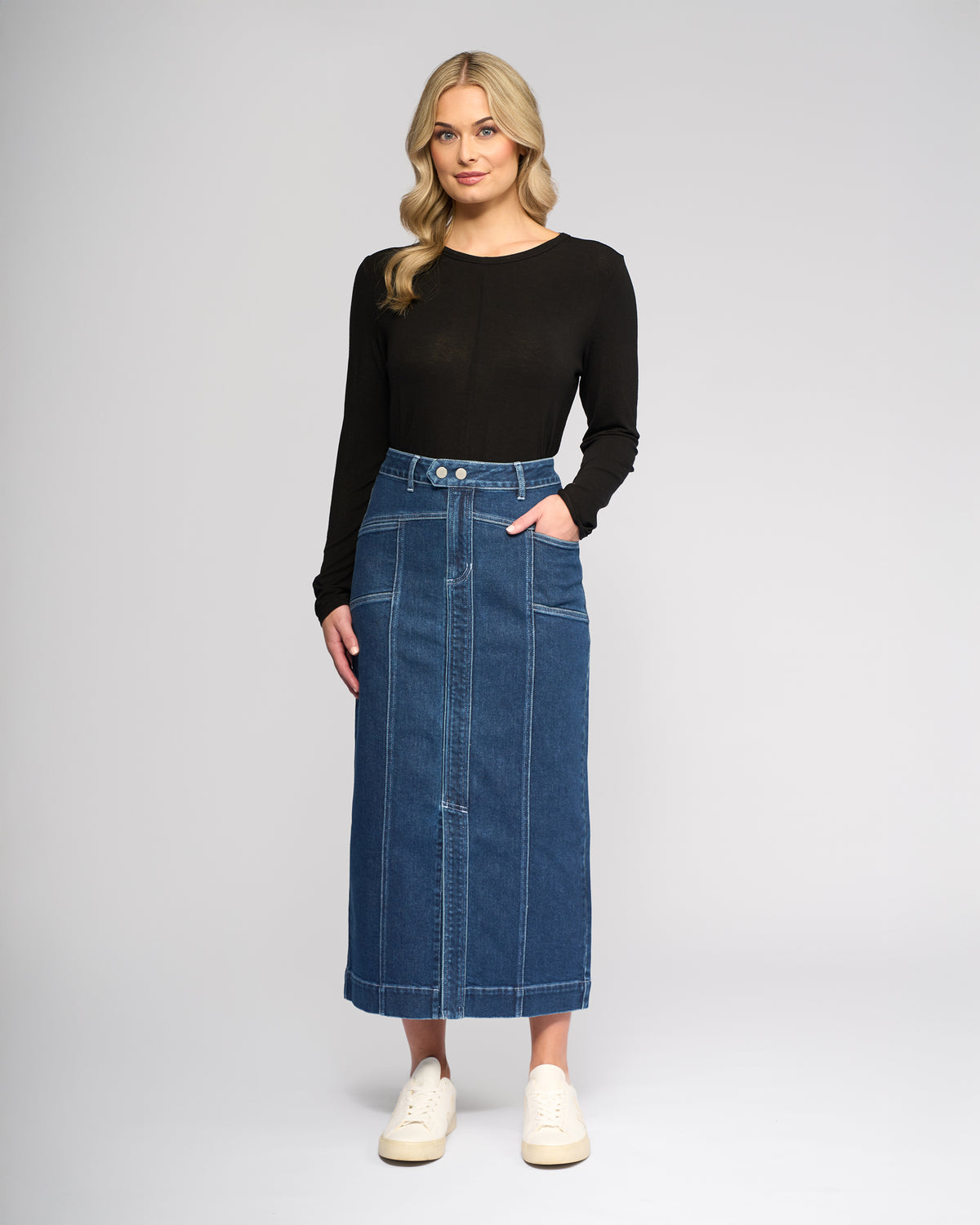 A woman with long blonde hair wears a black long-sleeve top, white sneakers, and the Vassalli Rook Skirt in Dark Blue—a high-waisted, panelled denim midi skirt with front seams and buttons—as she stands against a plain light background.