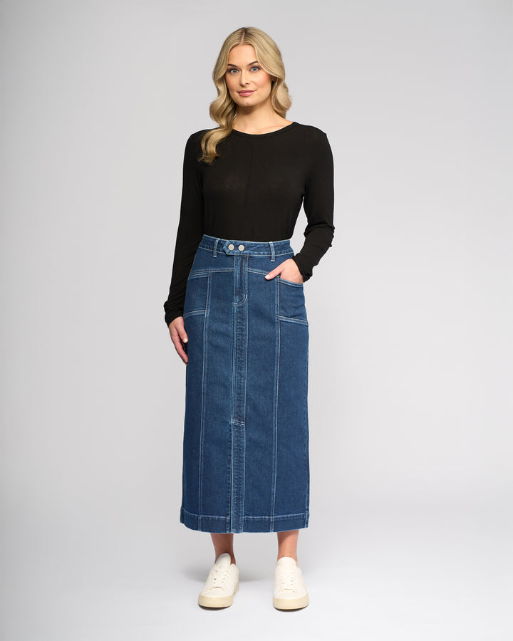 A woman with long blonde hair wears a black long-sleeve top, white sneakers, and the Vassalli Rook Skirt in Dark Blue—a high-waisted, panelled denim midi skirt with front seams and buttons—as she stands against a plain light background.