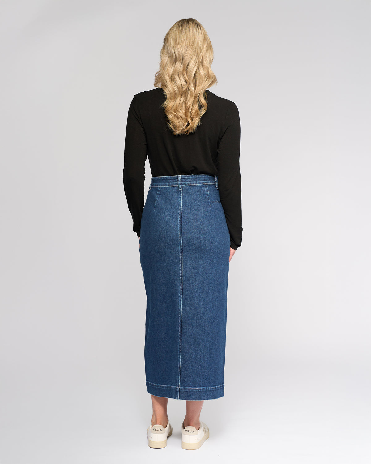 A woman with long, wavy blonde hair stands facing away, wearing a black long-sleeve top, white sneakers, and the Vassalli Rook Skirt - Dark Blue against a plain light background.