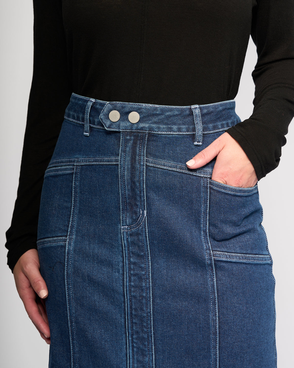 A person wearing a black long-sleeve top and the Vassalli Rook Skirt - Dark Blue, a high-waisted denim skirt featuring panelled design, two front buttons, visible stitching, and one hand in the skirt pocket.