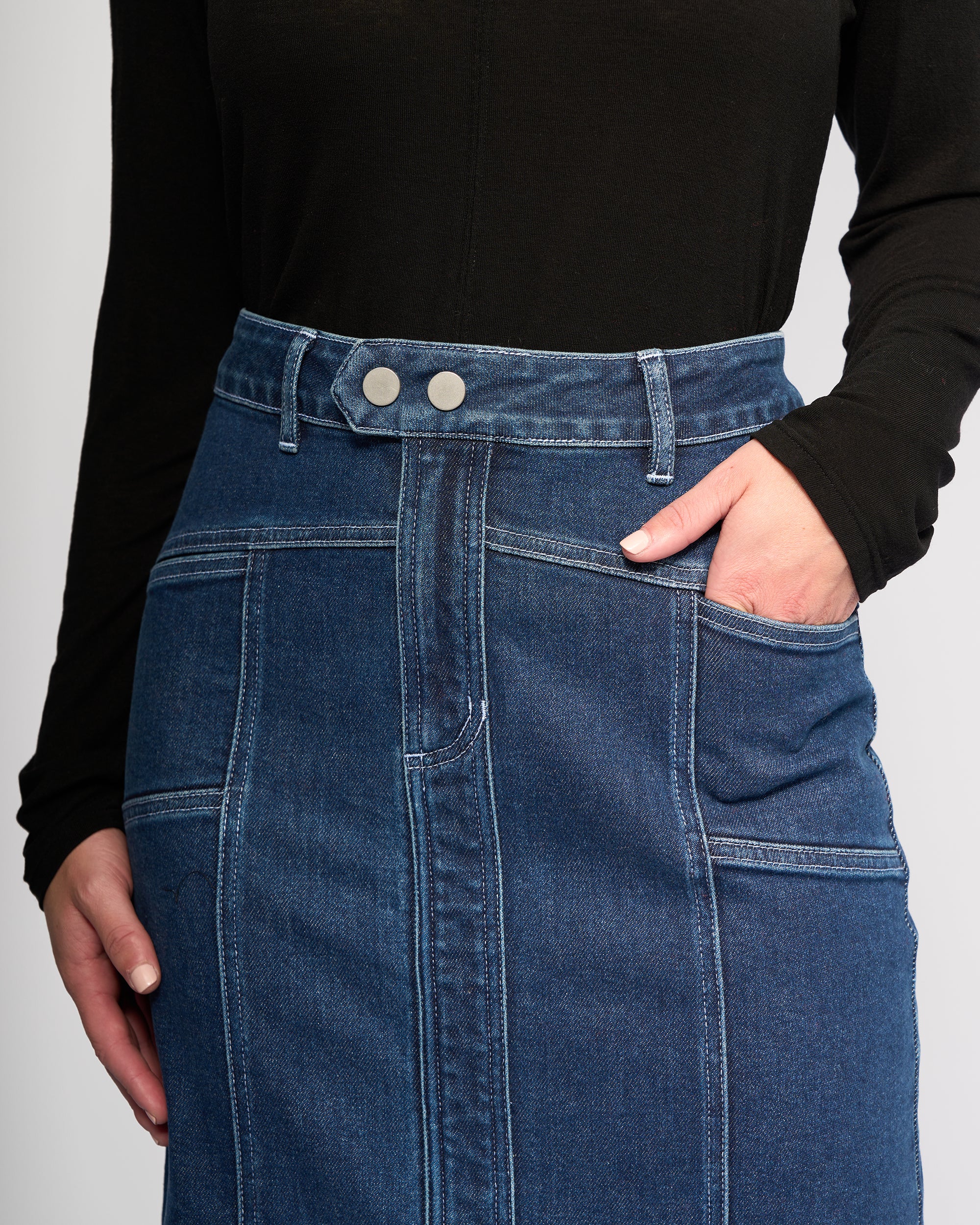 A person wearing a black long-sleeve top and the Vassalli Rook Skirt - Dark Blue, a high-waisted denim skirt featuring panelled design, two front buttons, visible stitching, and one hand in the skirt pocket.