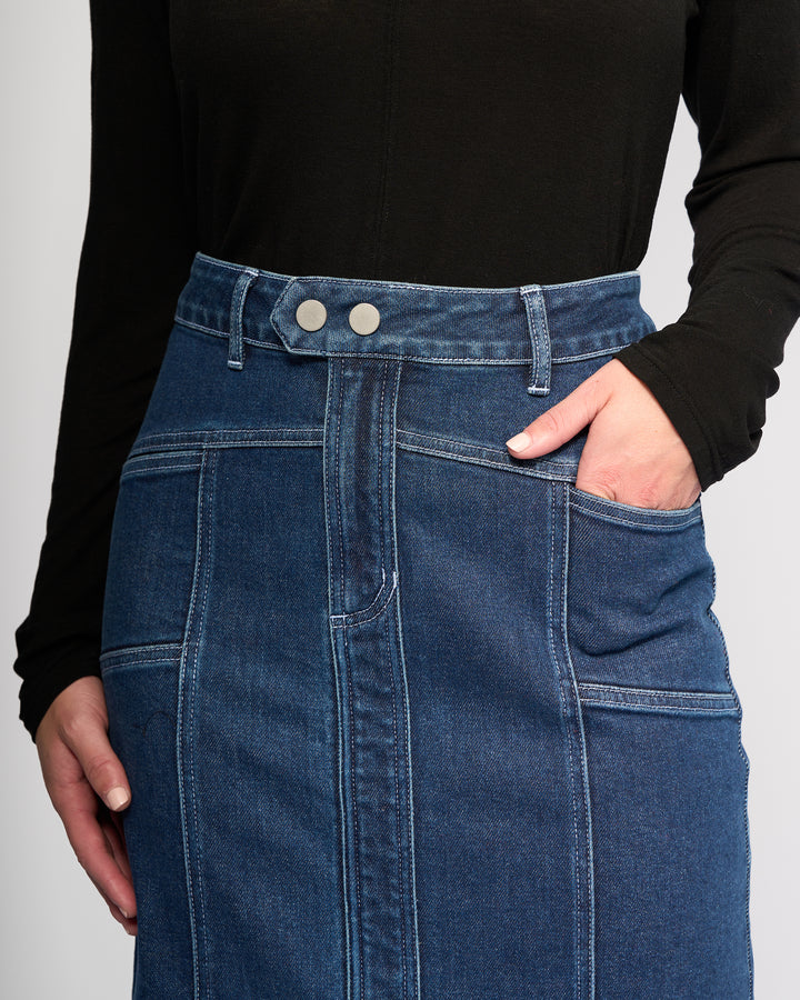 A person wearing a black long-sleeve top and the Vassalli Rook Skirt - Dark Blue, a high-waisted denim skirt featuring panelled design, two front buttons, visible stitching, and one hand in the skirt pocket.