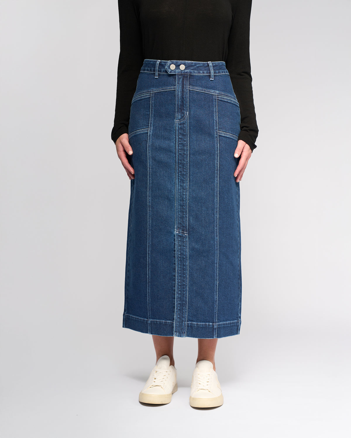 Someone stands against a plain light background wearing the Vassalli Rook Skirt - Dark Blue paired with a black long-sleeve top and white sneakers.
