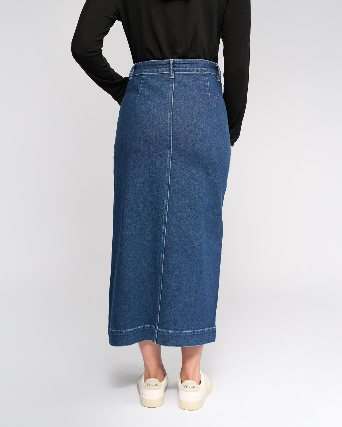 Someone wearing the Vassalli Rook Skirt in Dark Blue, a black long-sleeve top, and white sneakers with “VEJA” on the heels stands with hands behind their back, facing away from the camera.