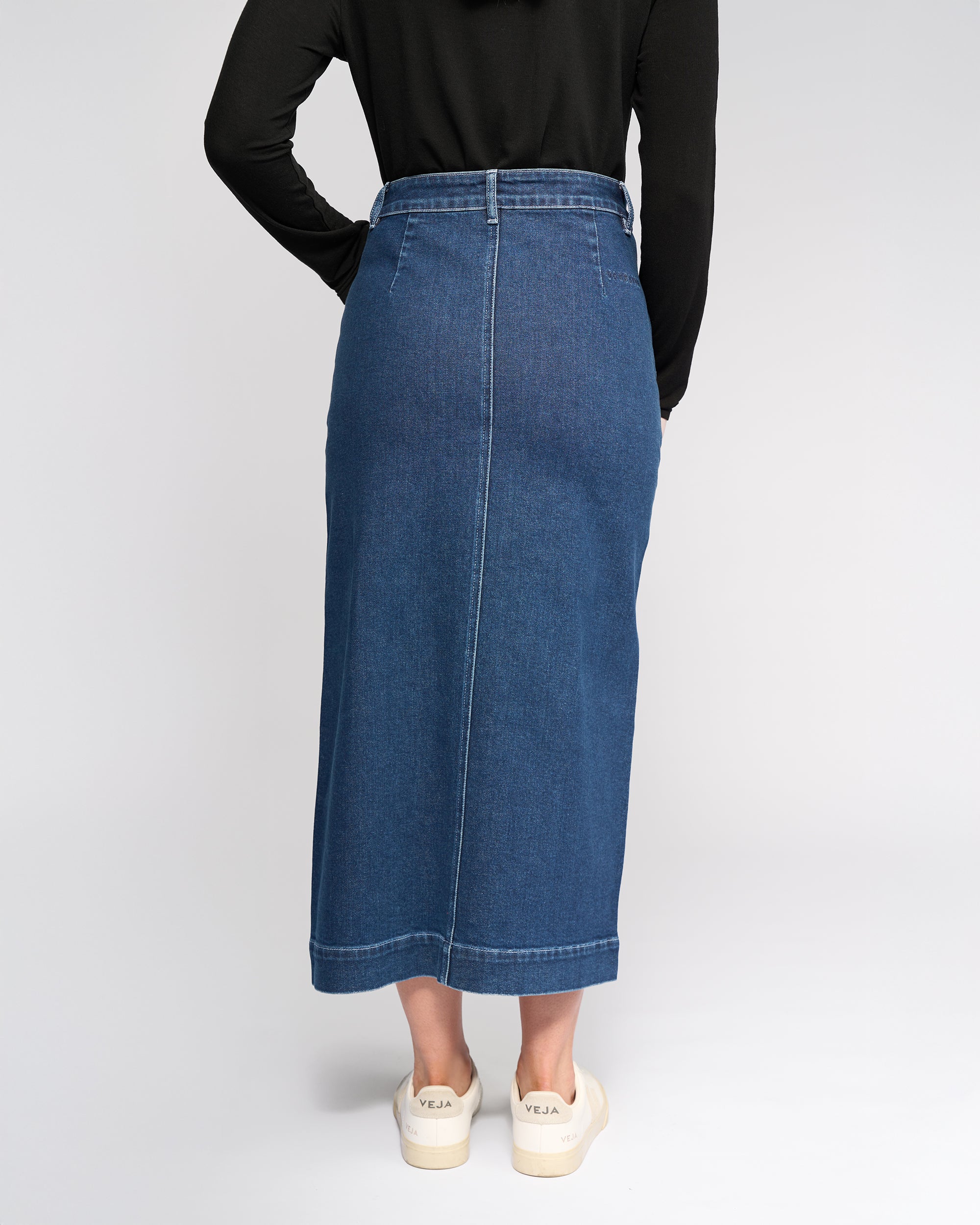 Someone wearing the Vassalli Rook Skirt in Dark Blue, a black long-sleeve top, and white sneakers with “VEJA” on the heels stands with hands behind their back, facing away from the camera.