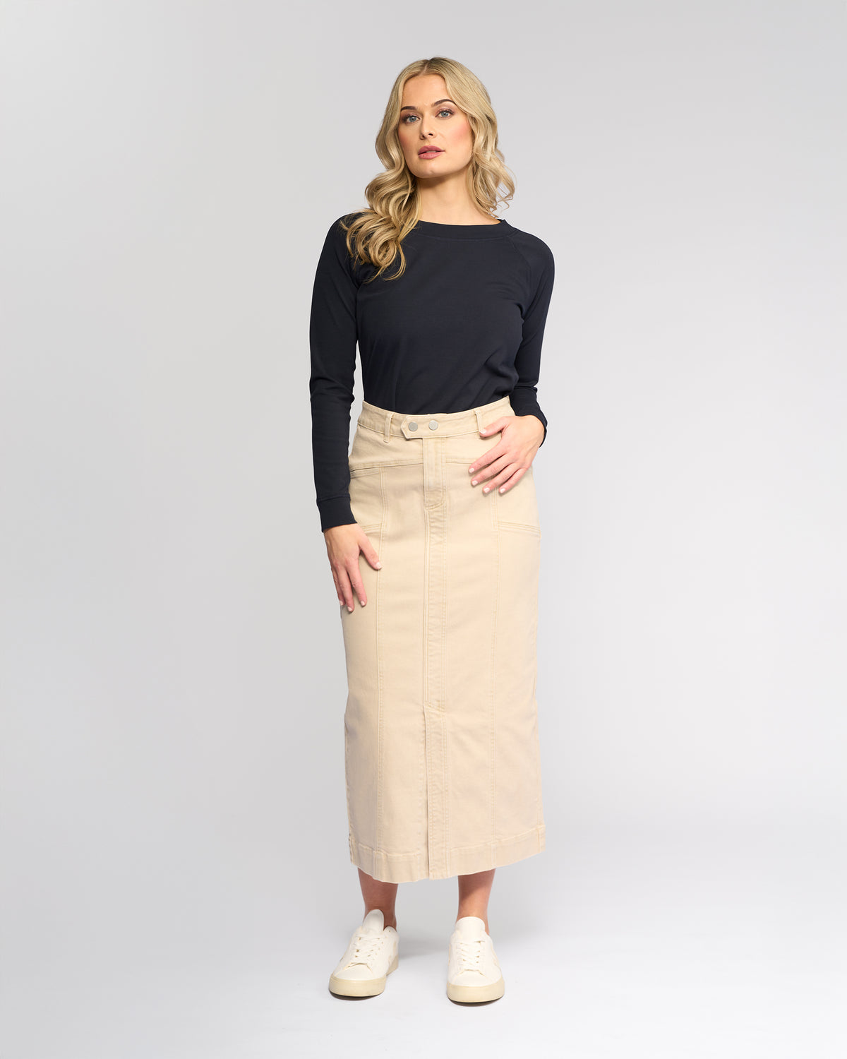 A woman with long blonde hair wears a black long-sleeve top, white sneakers, and the Vassalli Rook Skirt in Oat Denim. She stands against a plain light gray background, looking slightly to the side.