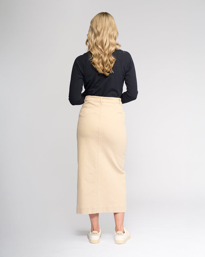 A woman with long wavy blonde hair, wearing a black long-sleeve top, the Vassalli Rook Skirt in oat denim, and white sneakers stands facing away from the camera against a plain light background.