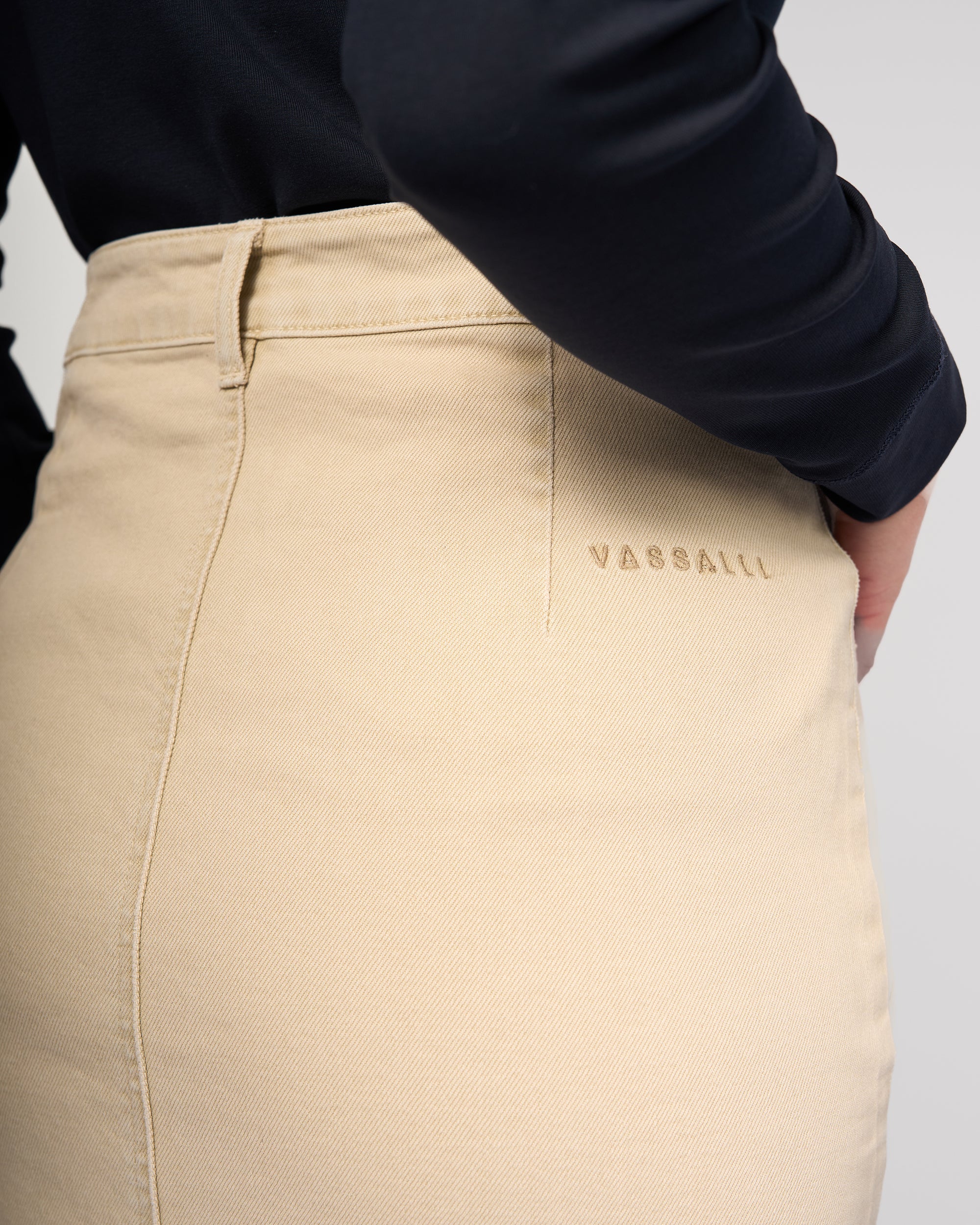 Close-up of a person wearing the Vassalli Rook Skirt - Oat Denim and a black long-sleeve top, showing the back with the VASSALLI logo embroidered near the waistband.