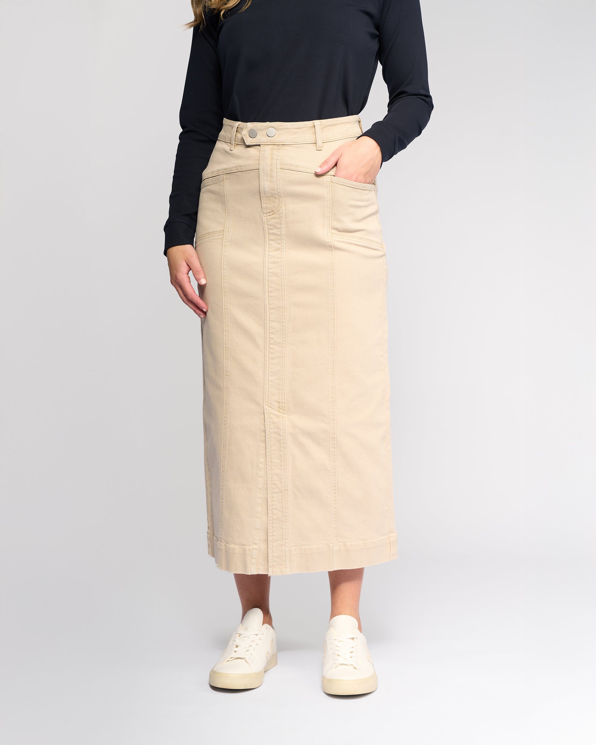 A person wearing a black long-sleeve top, white sneakers, and the Vassalli Rook Skirt in Oat Denim—a beige, panelled midi skirt with front pockets—stands against a light gray background. Their face is not visible.