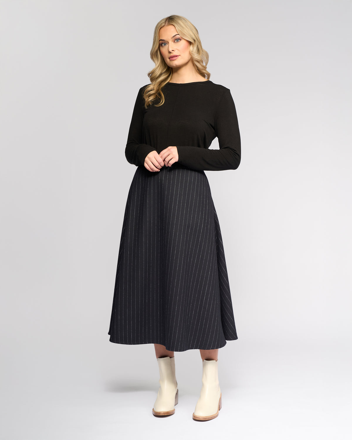 A woman with long, wavy blonde hair wears the Vassalli Gravity Skirt - Ink Stripe with a black long-sleeve top and white ankle boots, standing against a plain light gray background.