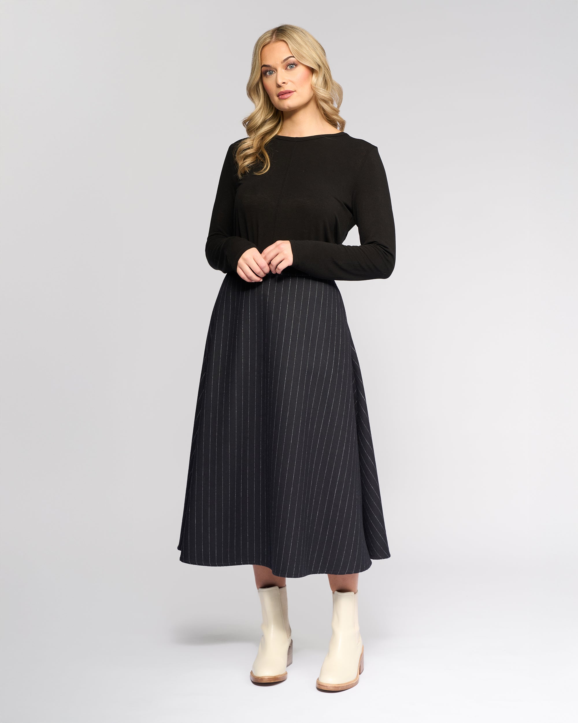 A woman with long, wavy blonde hair wears the Vassalli Gravity Skirt - Ink Stripe with a black long-sleeve top and white ankle boots, standing against a plain light gray background.